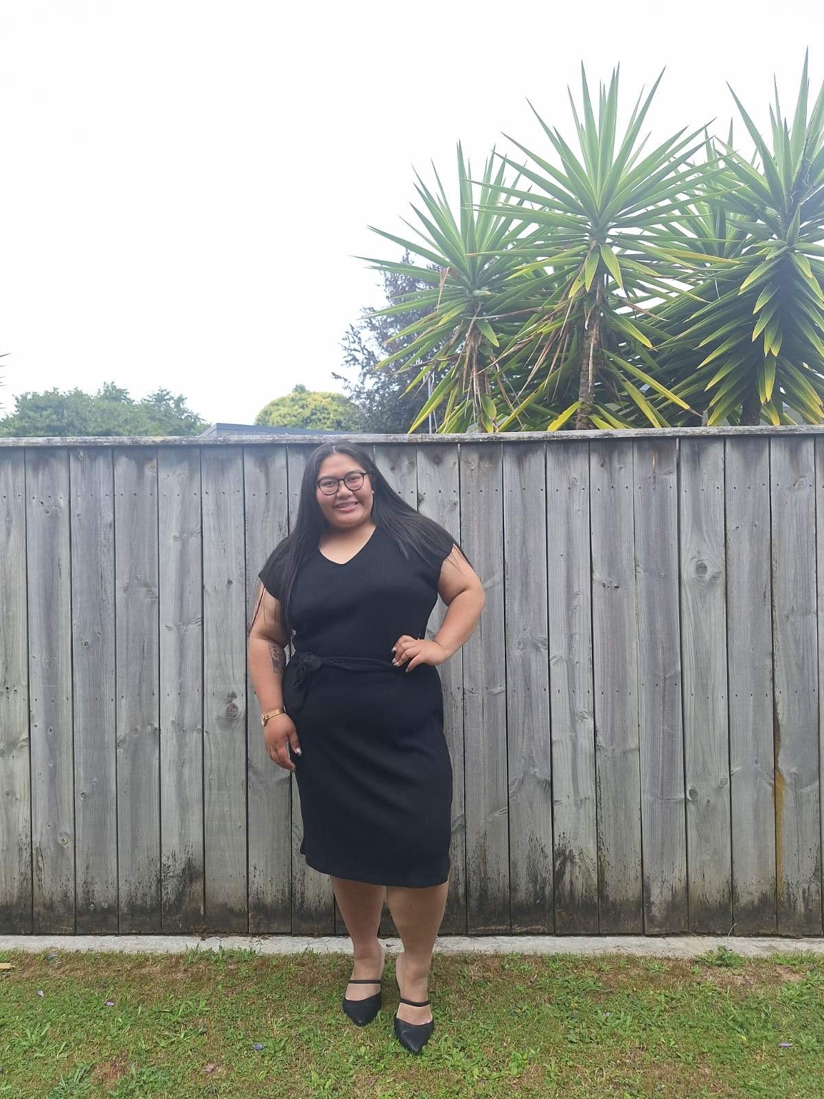 A woman with long black hair and glasses, wearing a black dress and heels, stands in front of a wooden fence with green plants and trees in the background. The sky is overcast.