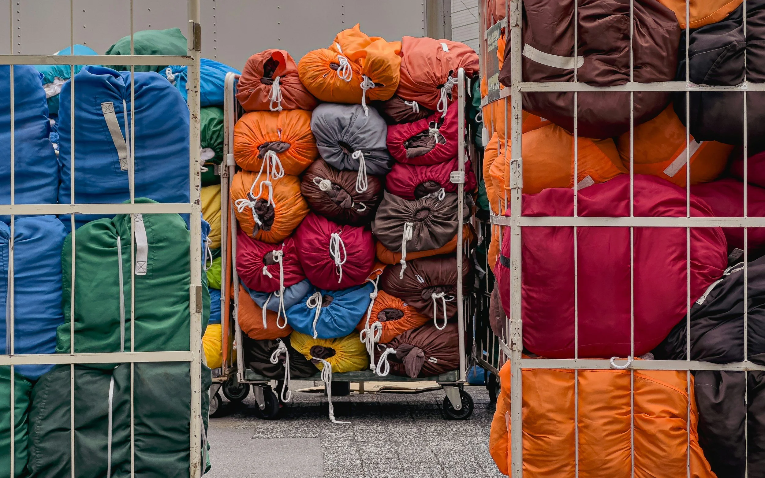 A shopping cart filled with colorful sleeping bags in shades of blue, green, orange, red, brown, and gray is seen at a store or warehouse, with metal shelving around it.