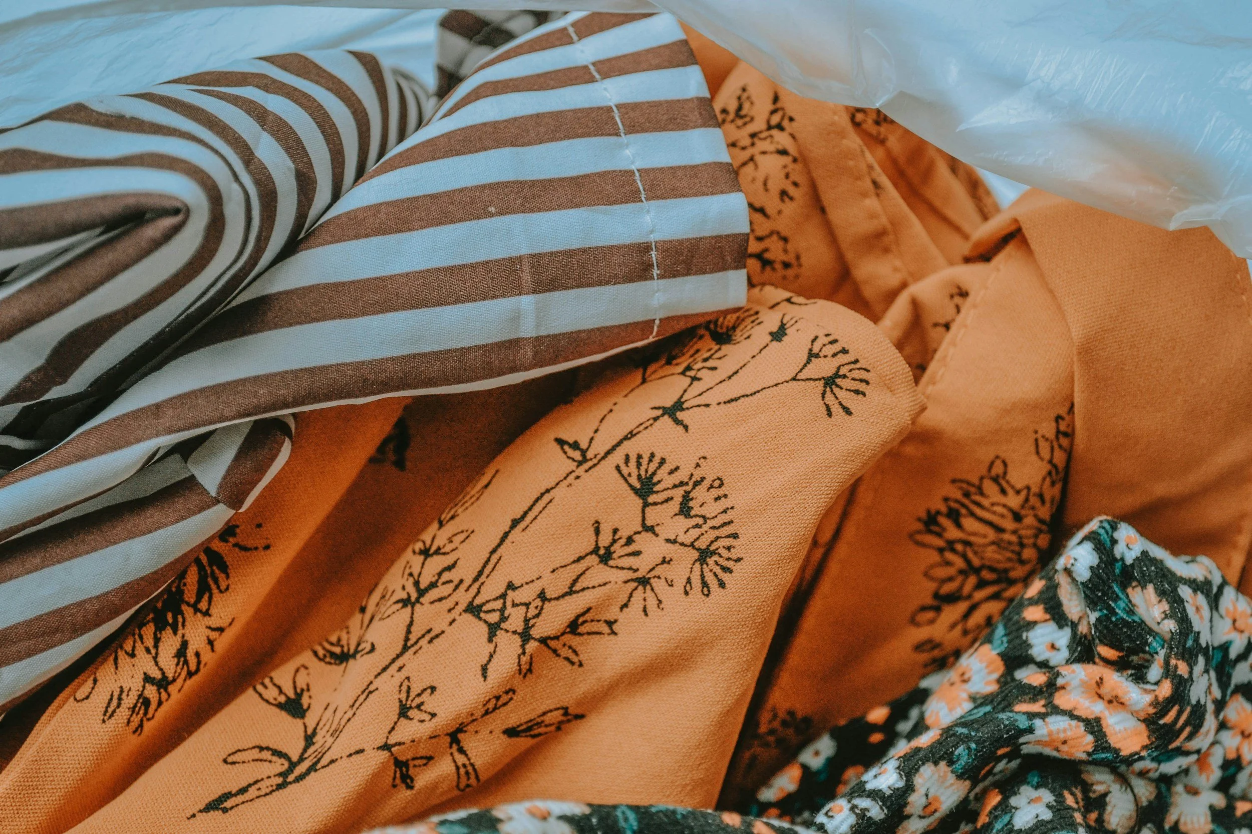 Close-up of a pile of fabric with various patterns, including orange with black floral and branch designs, striped brown and white, and black and orange floral patterns.