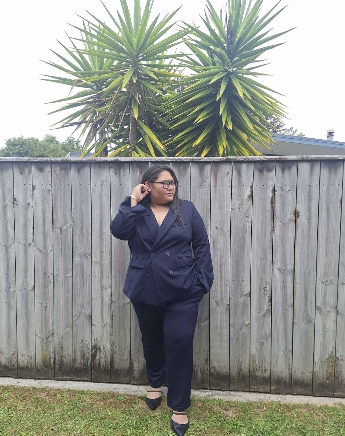 A woman in a navy suit standing against a wooden fence with tall green spiky plants behind her.