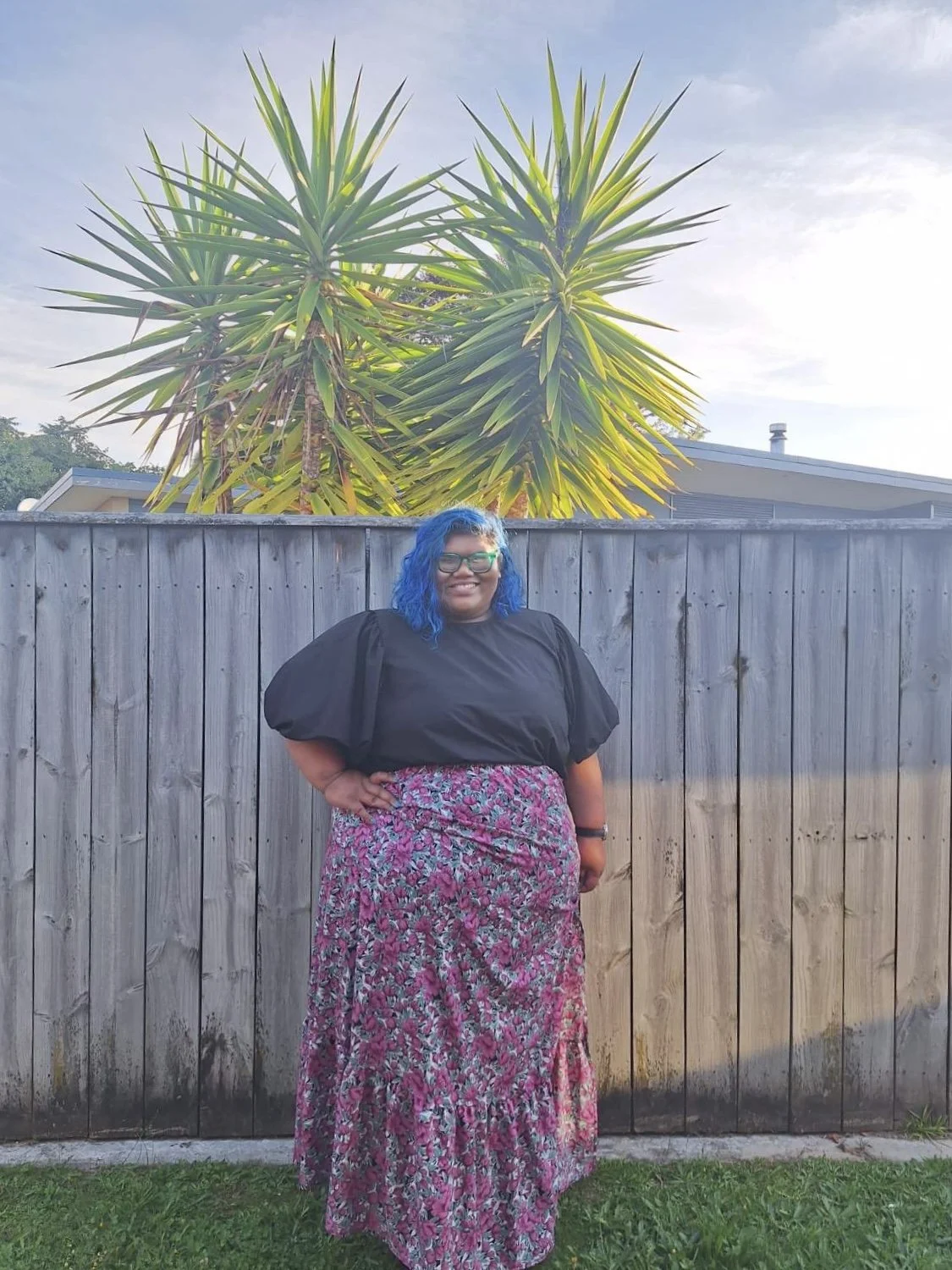 A woman with blue hair and glasses standing outdoors in front of a wooden fence and a large green plant with pointed leaves, wearing a black top and a long pink floral skirt.
