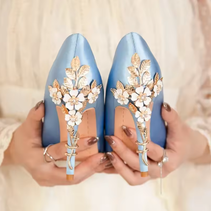 Close-up of a pair of light blue high-heeled shoes with white floral embroidery and gold accents, held by person's hands.