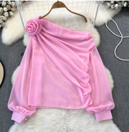 Pink chiffon dress with a floral rosette detail on the shoulder, draped fabric, and gathered cuffs, displayed on a hanger on a white furry surface.
