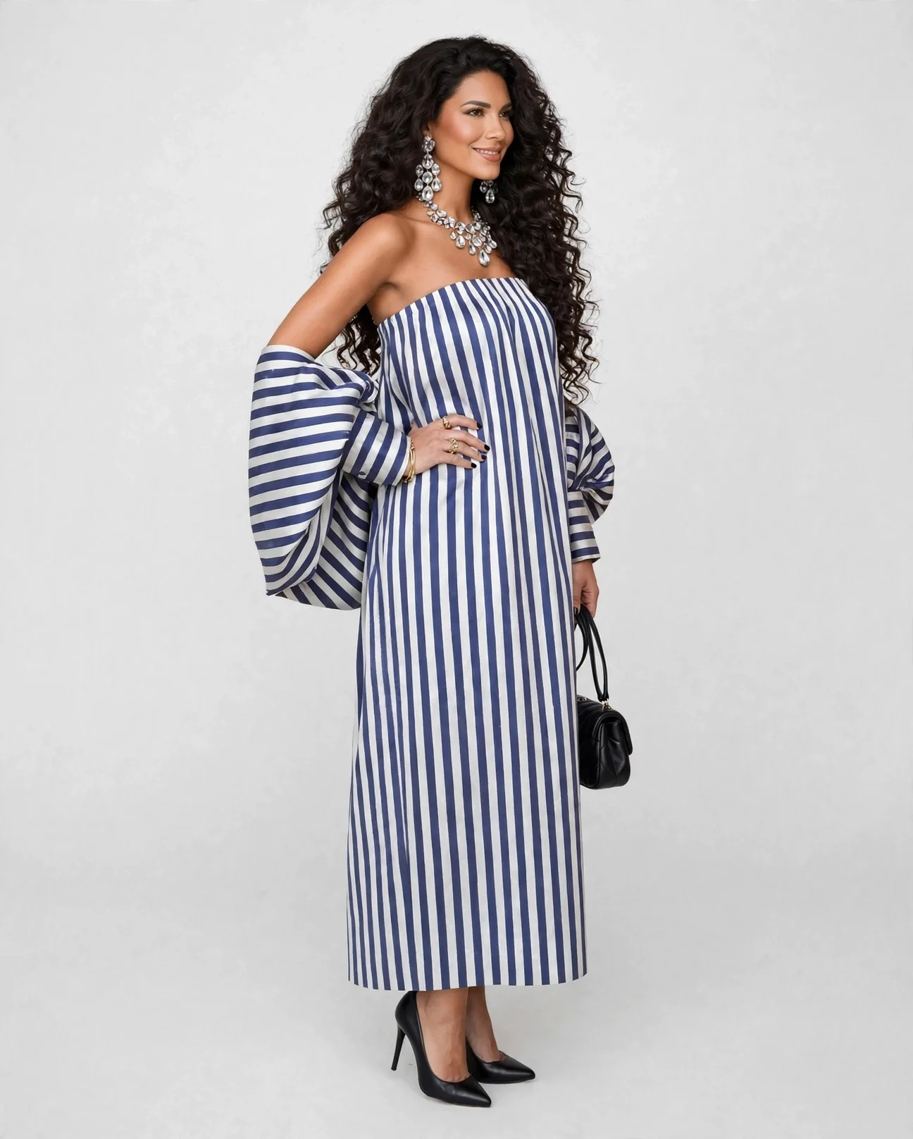 Woman with long curly dark hair wearing a strapless blue and white striped dress, matching striped sleeves, black high heels, large statement jewelry, and holding a small black handbag against a plain white background.