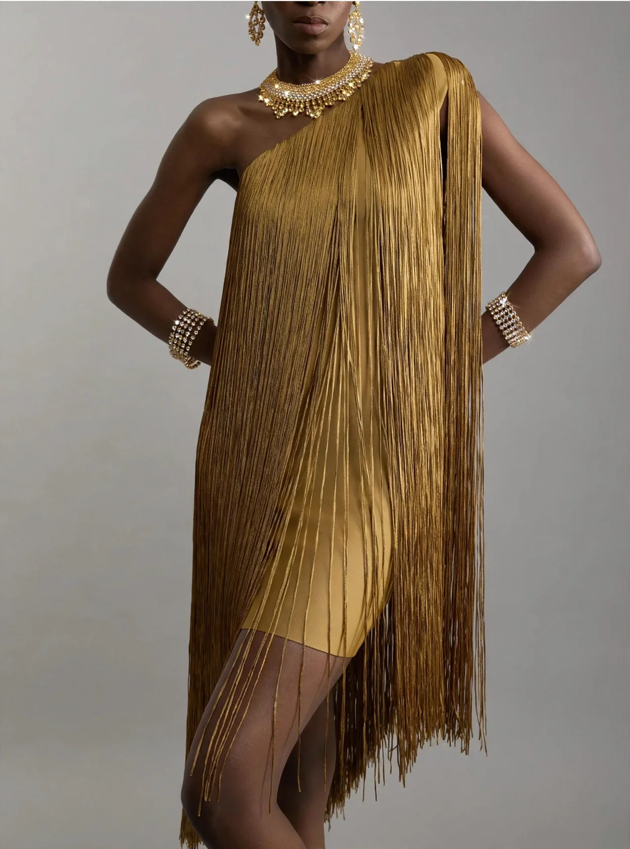 A woman wearing a gold pleated dress with fringe details, gold jewelry including necklaces, earrings, and bracelets, standing against a plain background.