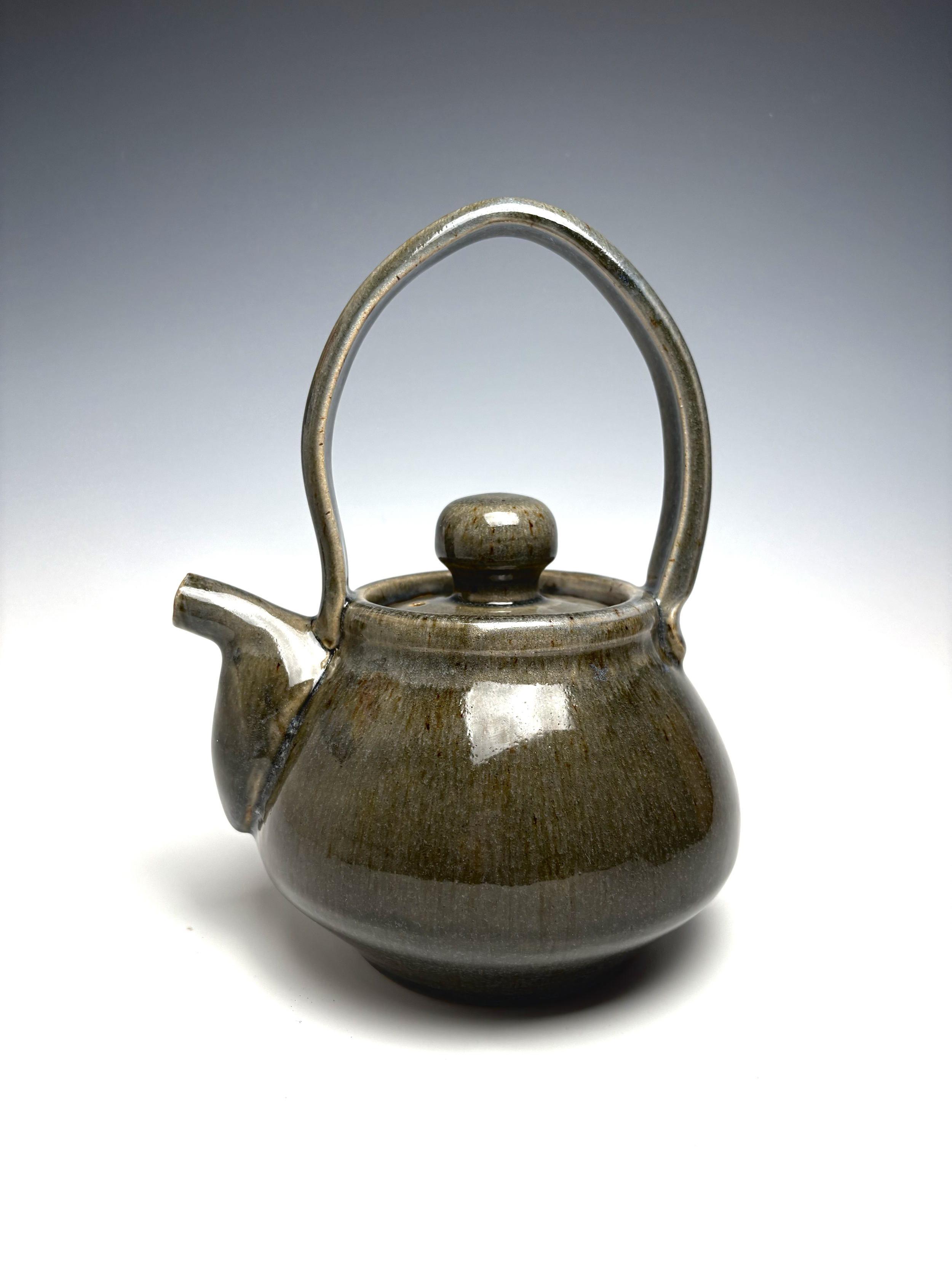 A ceramic teapot with a rounded body, a curved spout, a handle that forms an arch above the lid, and a small knob on the lid, set against a plain background.