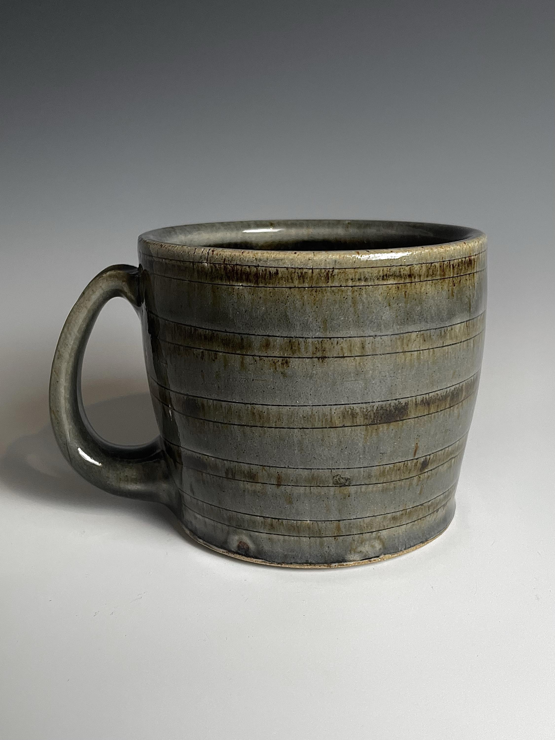 A ceramic mug with horizontal ridges and a handle on the left side, featuring a glazed, textured surface with streaks of brown and gray color.