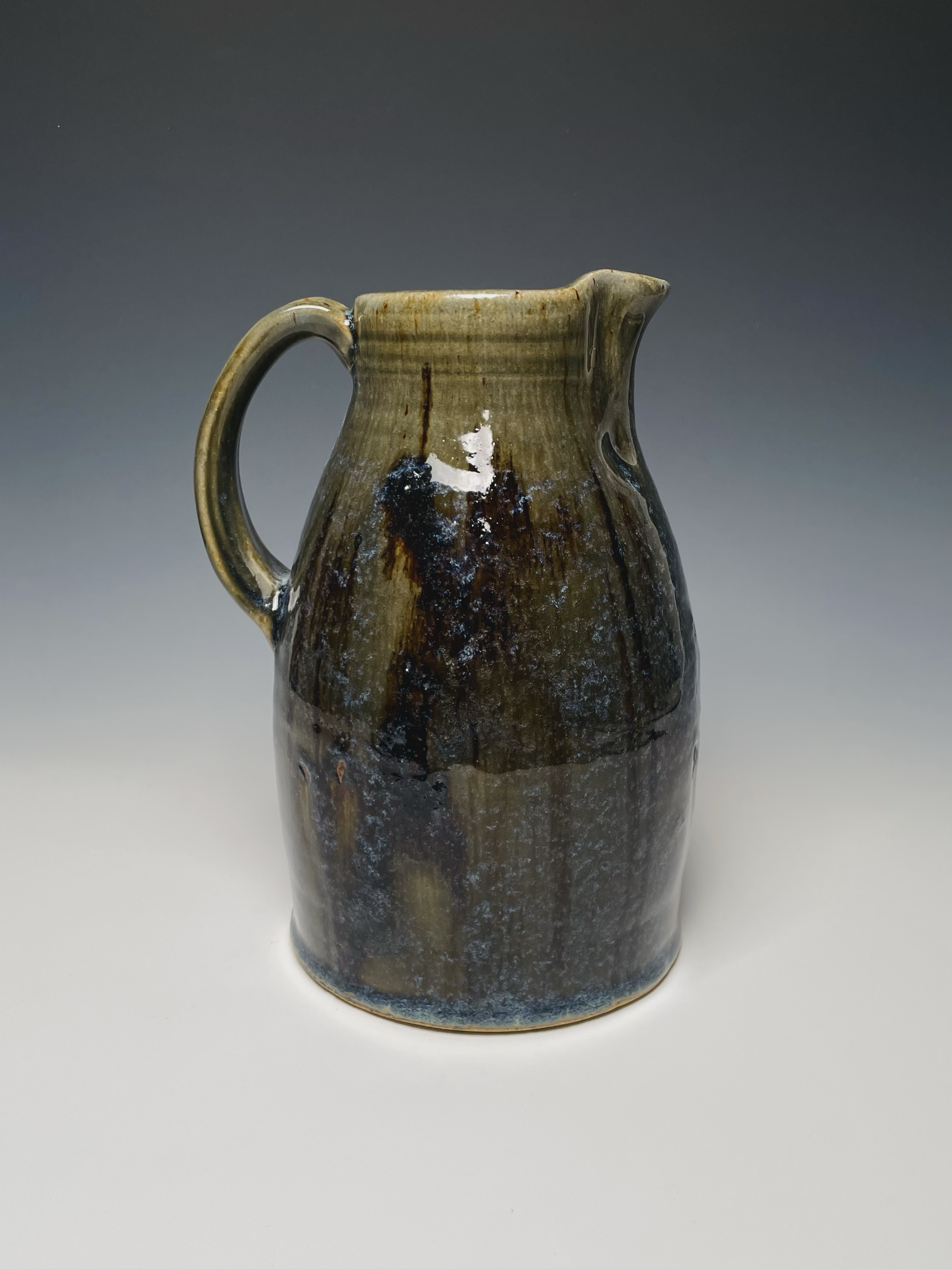 A ceramic pitcher with a handle and spout, glazed in earthy tones with a mottled, textured surface, set against a plain gray background.