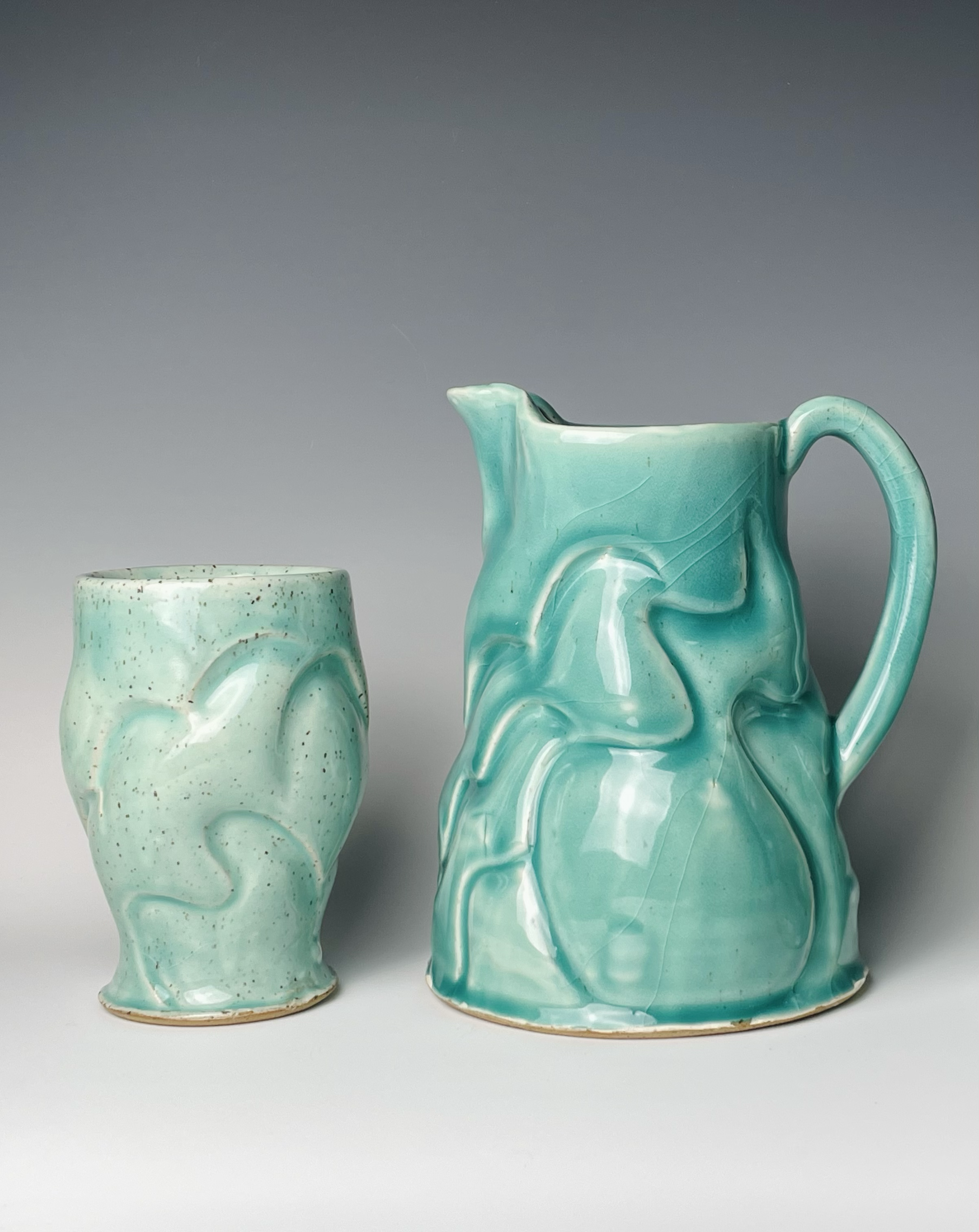 A pair of ceramic turquoise-colored vases with swirl and wave patterns, one short and round, the other tall with a spout and handle, set against a plain gray background.