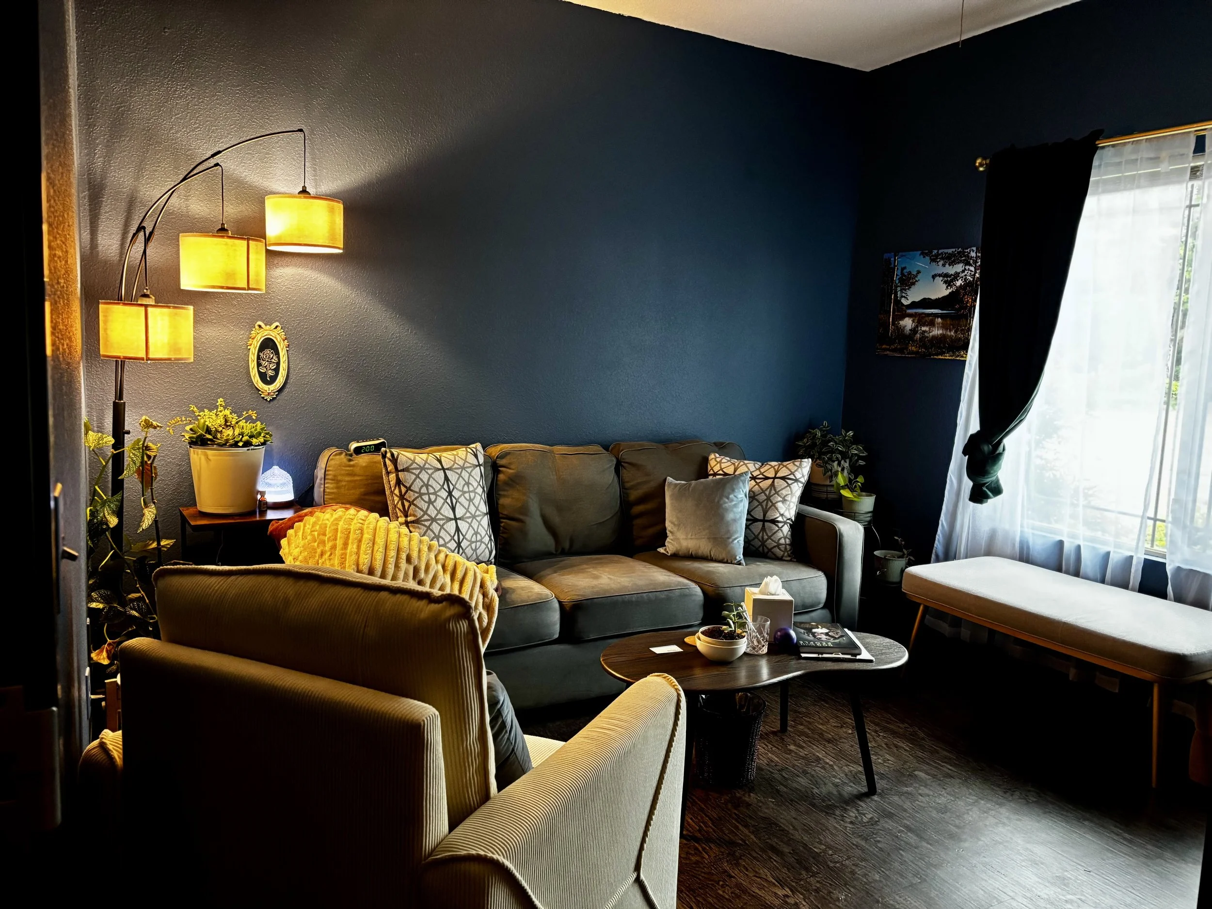 A cozy living room with dark blue walls decorated with a small mirror and a landscape painting. There are several sofas, one striped and others in solid colors, with cushions. A wooden coffee table holds a bowl, a magazine, and a notebook. A floor lamp with three yellow lampshades illuminates the space. A large window with sheer white curtains and dark drapes allows natural light into the room. There are also potted plants and decorative items, creating a warm, inviting atmosphere.