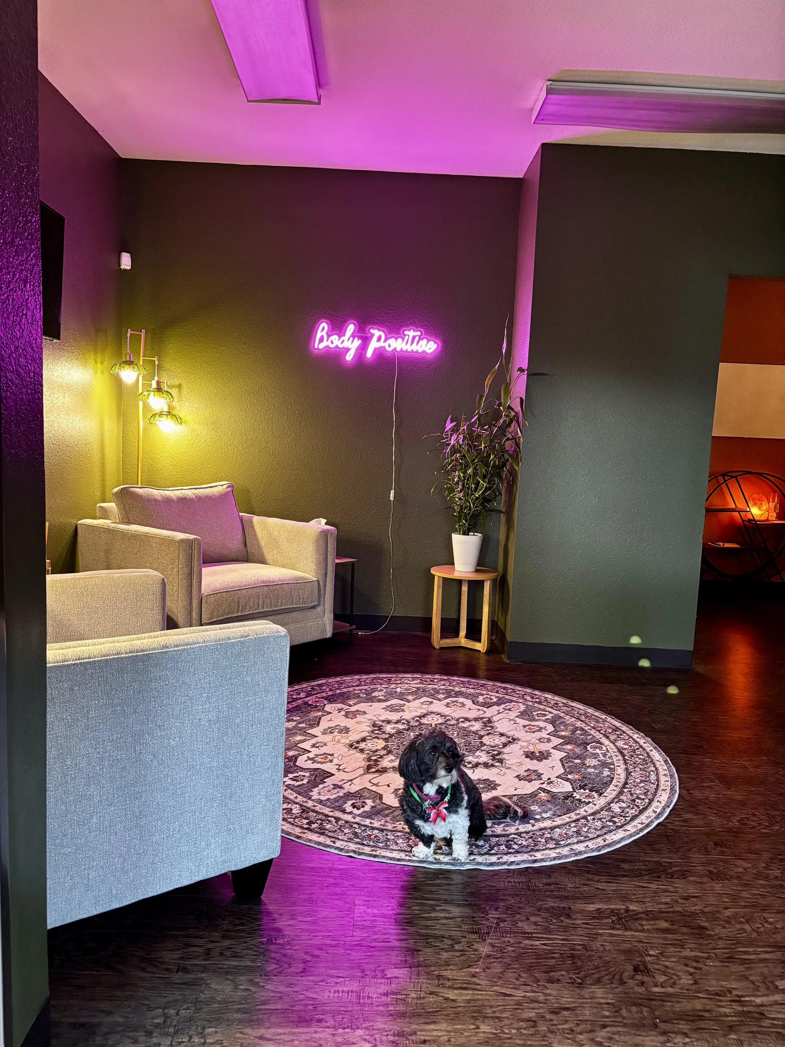 A cozy lounge with a black and white poodle puppy sitting on a patterned rug, green, pink, and purple lighting, a beige armchair, a neon sign reading 'Body Positivity', a potted plant, and modern decor.