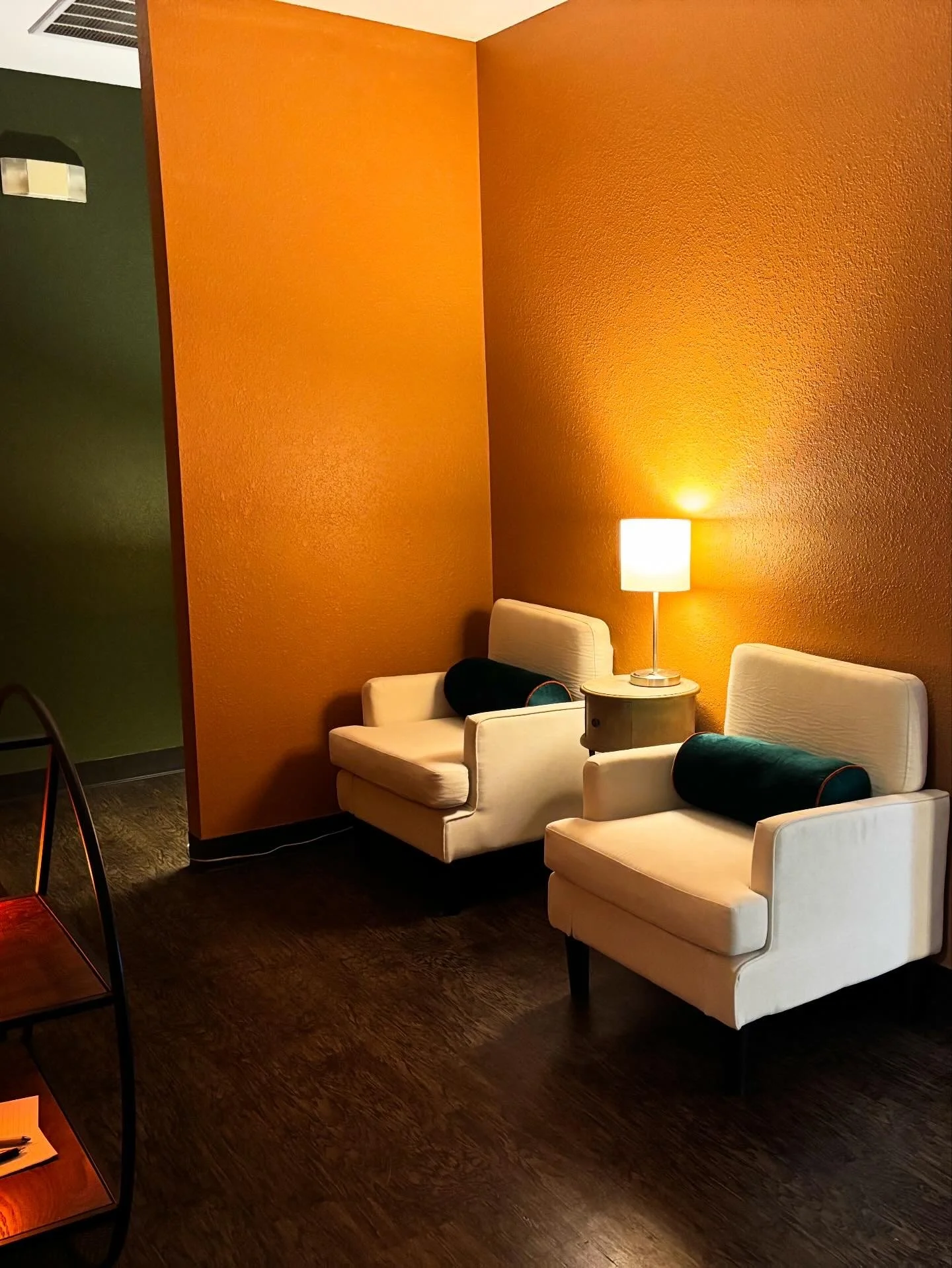 Living room corner with two cream armchairs, a table lamp, and a small side table, against orange and green walls, with dark wood flooring.