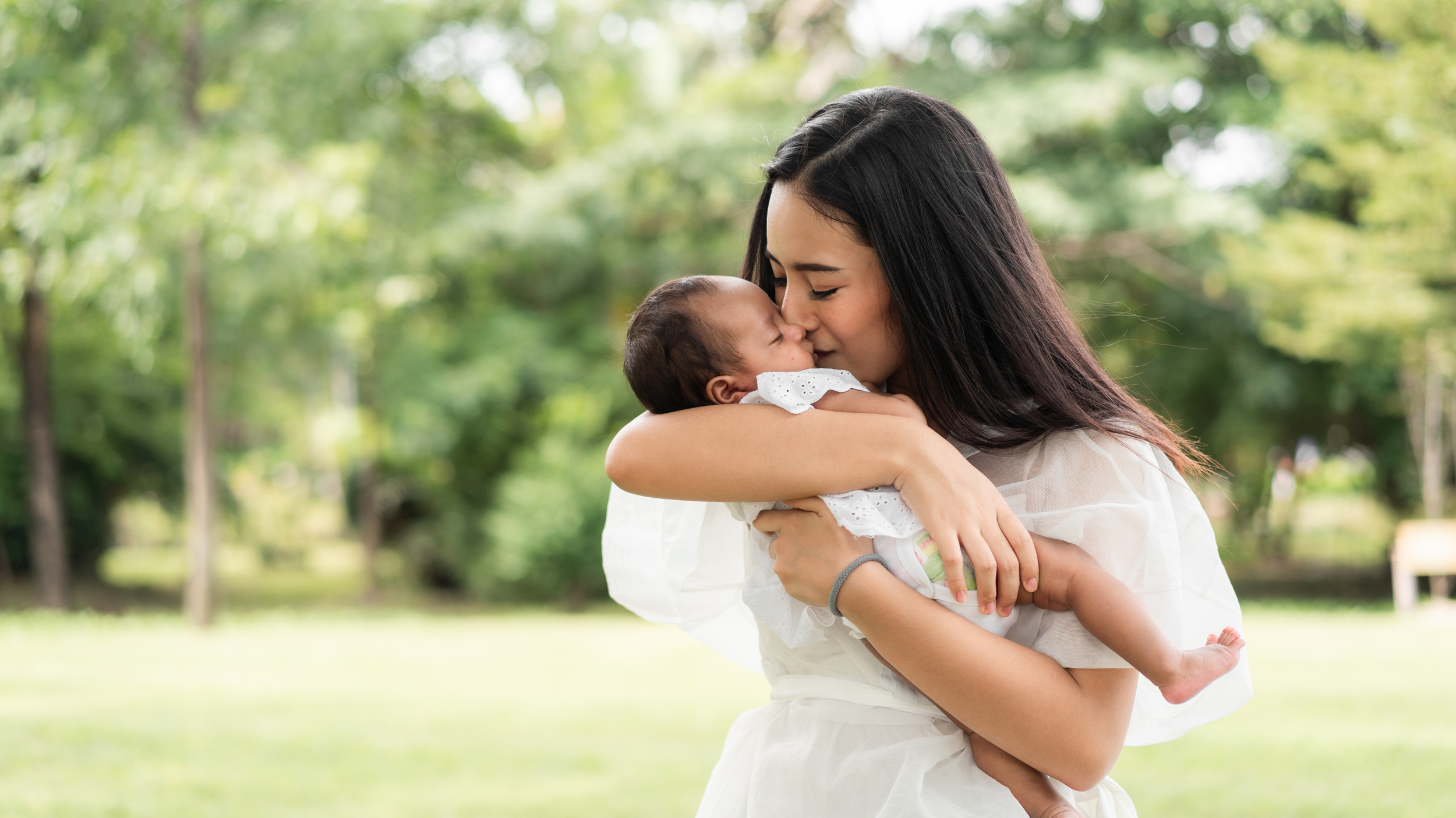 A woman with long dark hair holding a baby close, both touching noses in a tender moment outdoors with green trees in the background.