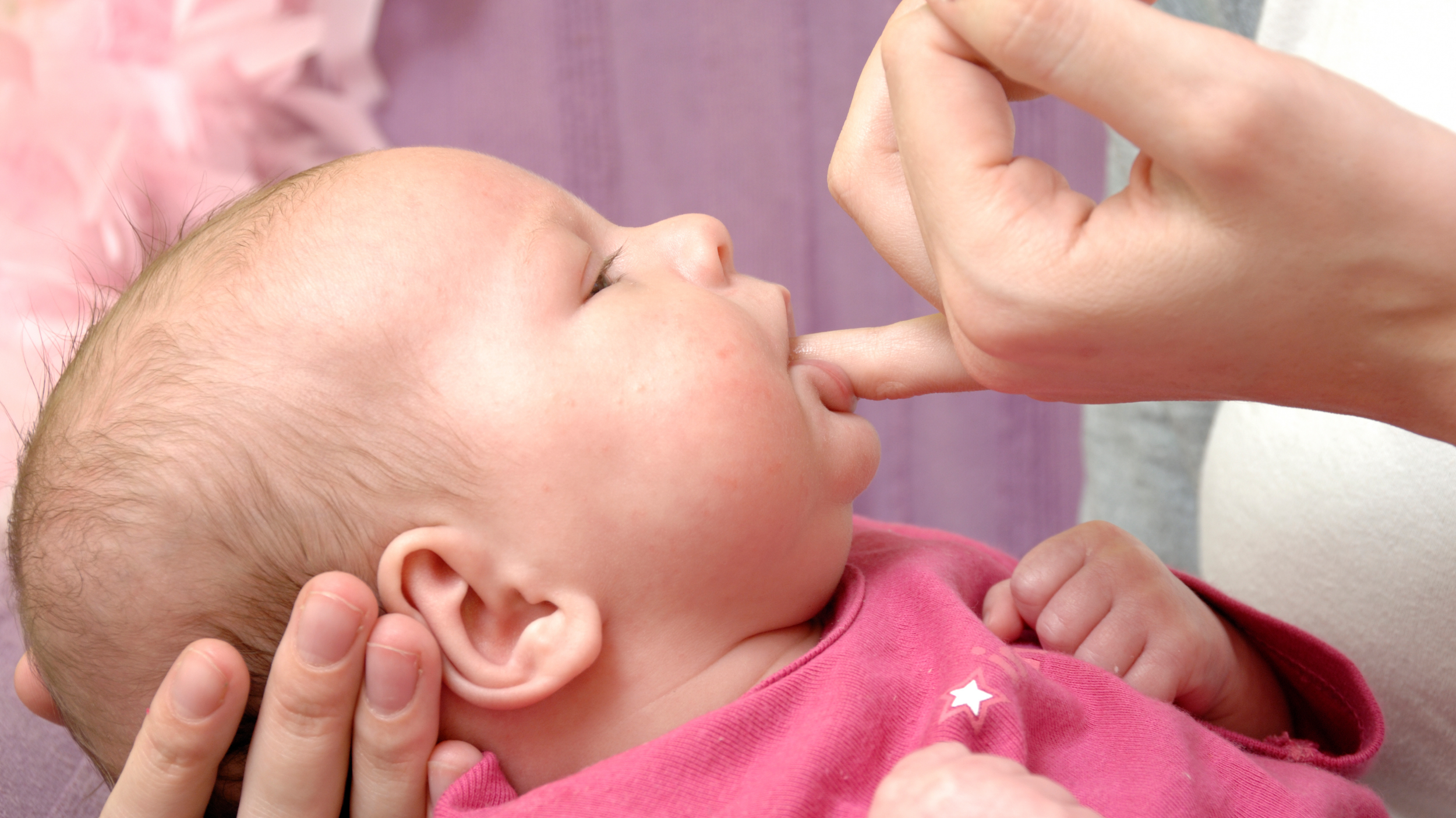 A baby with light skin and blond hair is lying down while an adult gently touches their nose with a finger. The baby is wearing a pink shirt with white star patterns and appears to be looking at the adult's hand.