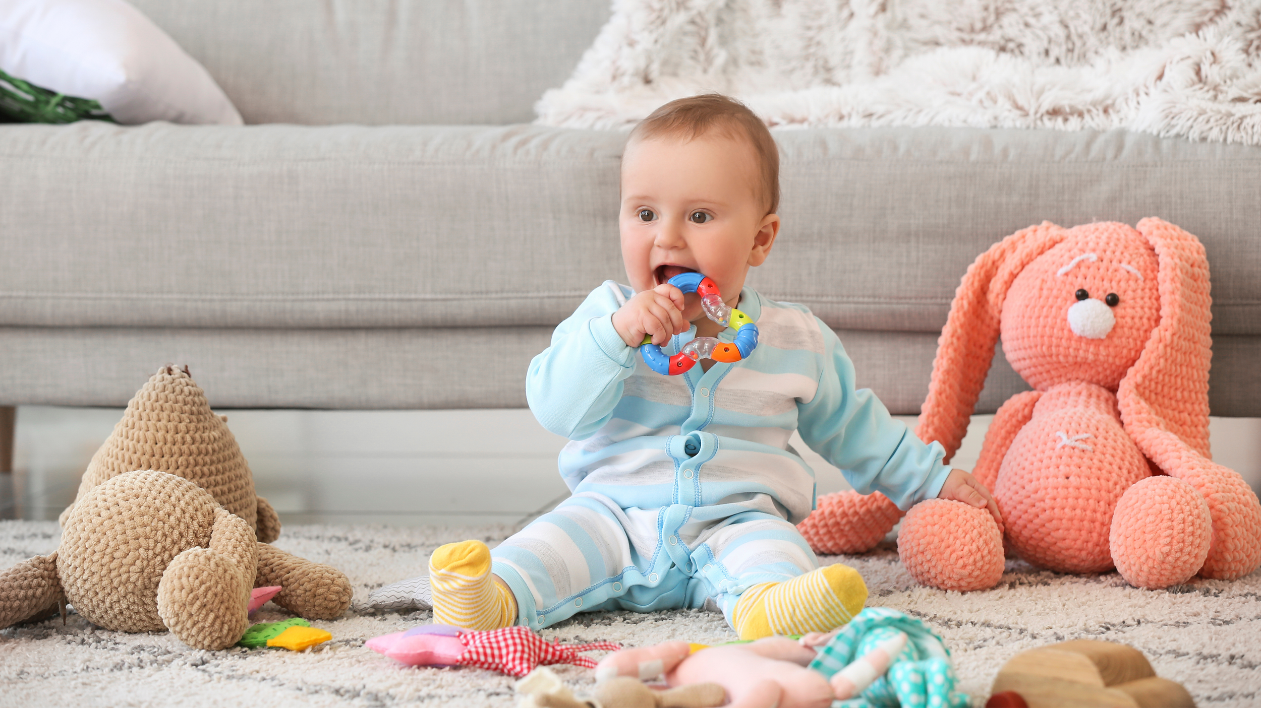 A baby sitting on a rug with toys and stuffed animals, holding a colorful teething ring in mouth, in a cozy living room with a gray sofa and decorative pillows.