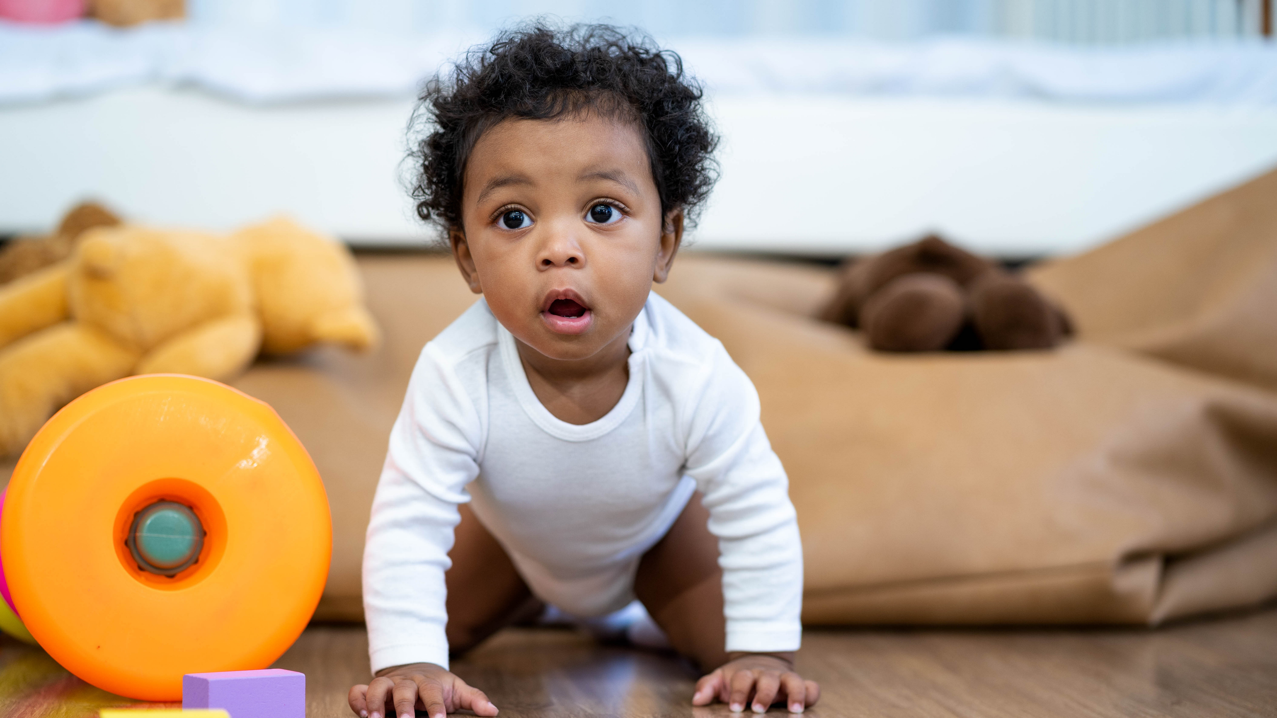 A young child with curly hair crawling on the floor, surrounded by colorful toys in a playroom.