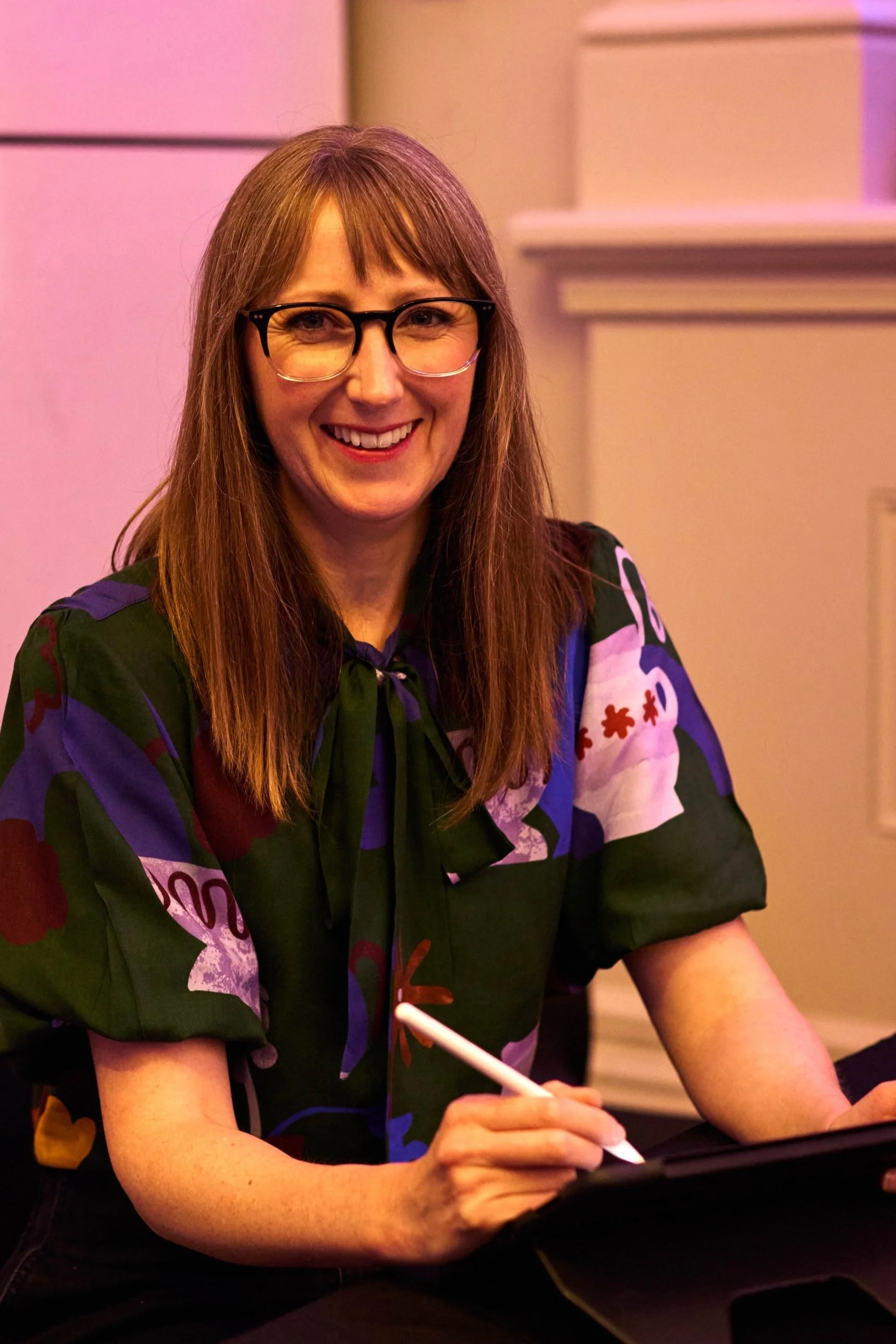 A smiling woman wearing glasses with straight brown hair, dressed in a colorful patterned blouse, sitting at a table with a digital tablet and stylus.