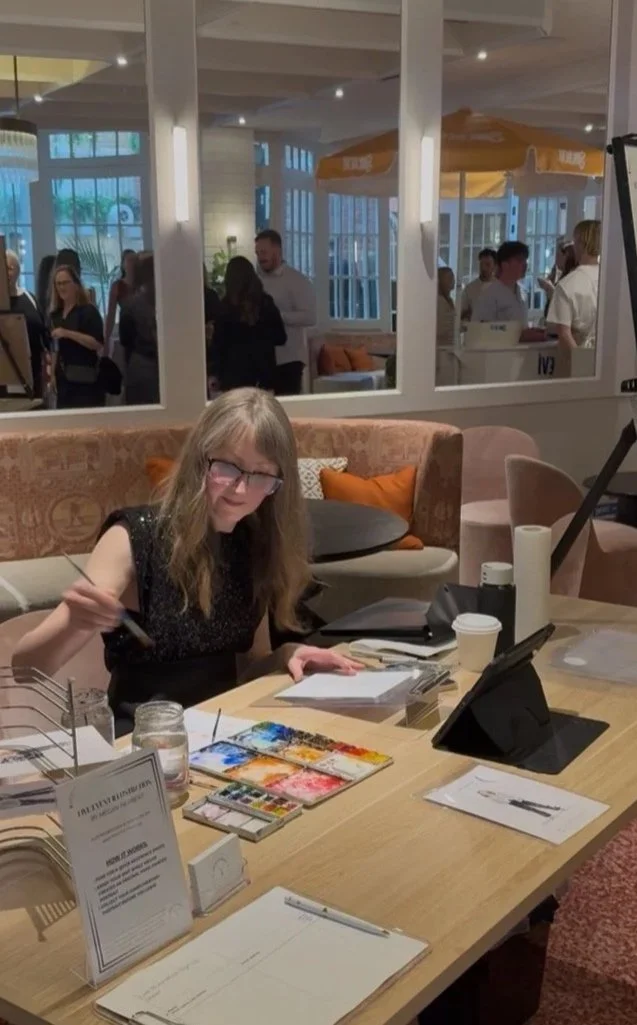 Megan Herbert, a woman with long blonde hair, and the Mornington Peninsula's number one wedding and event illustrator, is smiling while painting at a crowded brand activation event for the Continental Sorrento's CeeCee's restaurant.