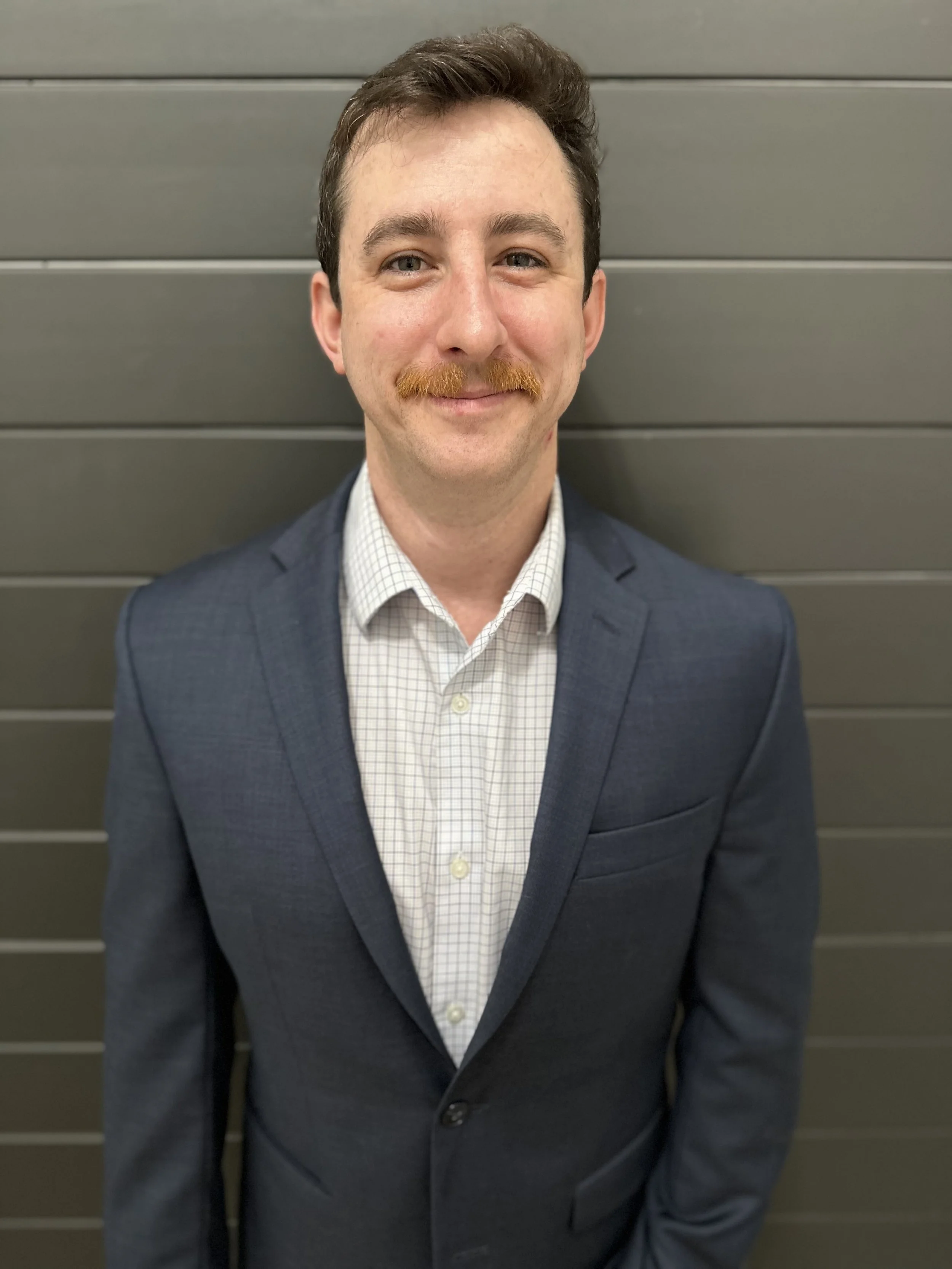 A man in a navy suit and checkered white shirt standing in front of a gray horizontal panel background.