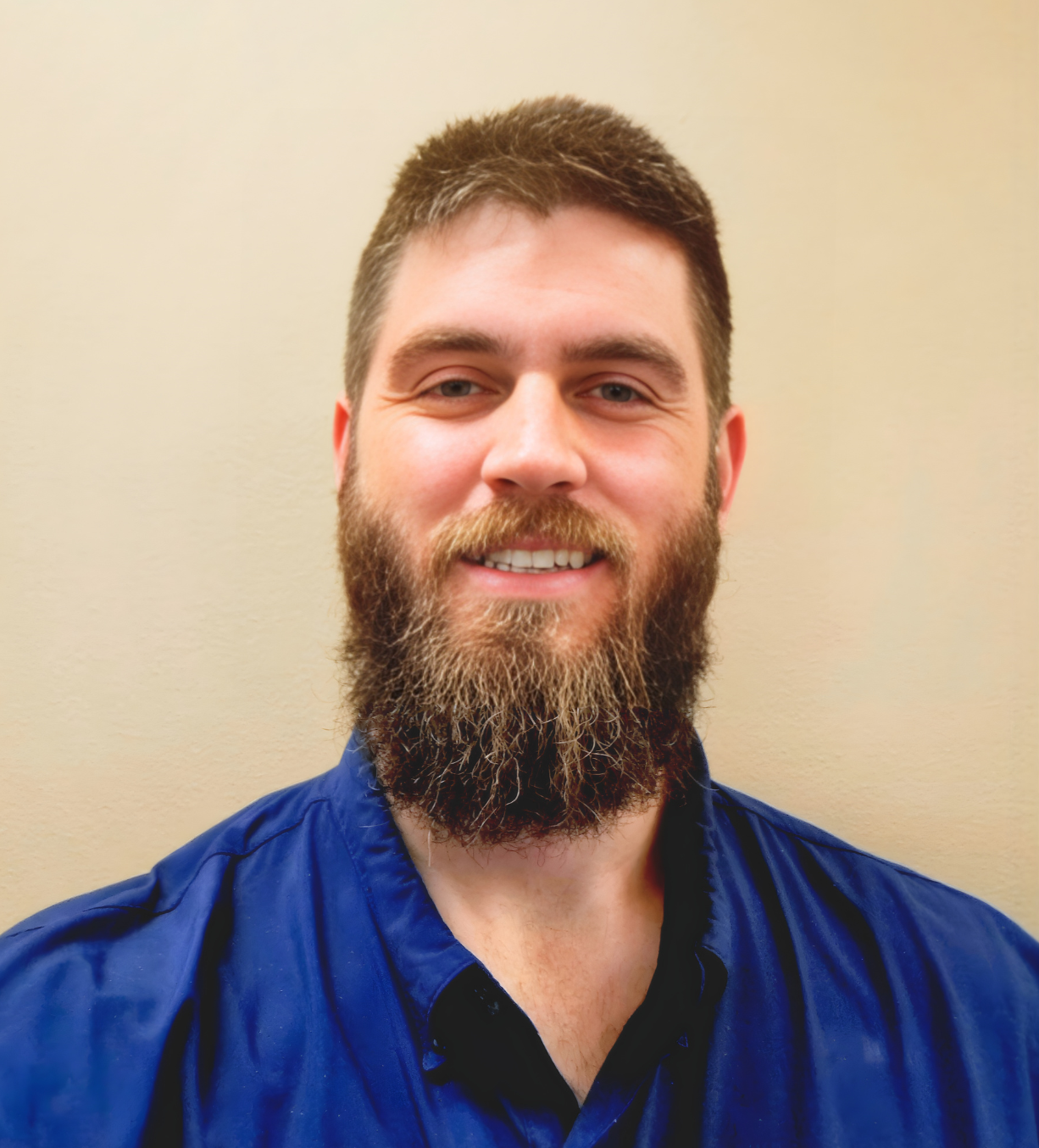 A smiling man with a beard and short brown hair wearing a blue collared shirt standing against a beige wall.