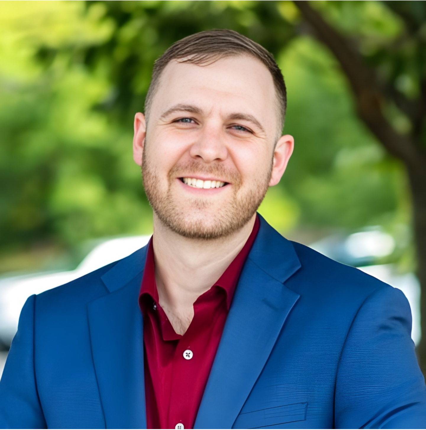 A smiling man with light skin, brown hair, and a beard, wearing a blue blazer and maroon shirt, standing outdoors with green trees in the background.