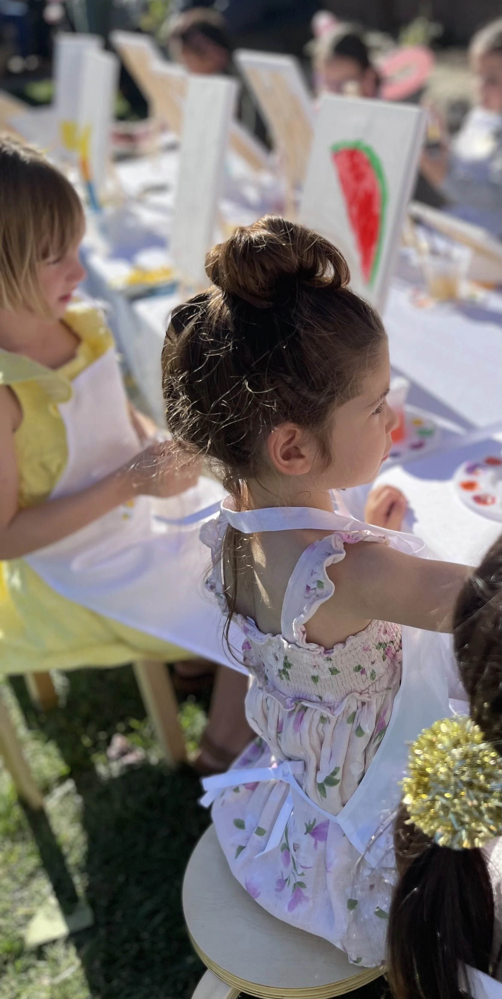 Children painting on canvas at an outdoor art party, sitting at a long table with art supplies and artworks displayed.