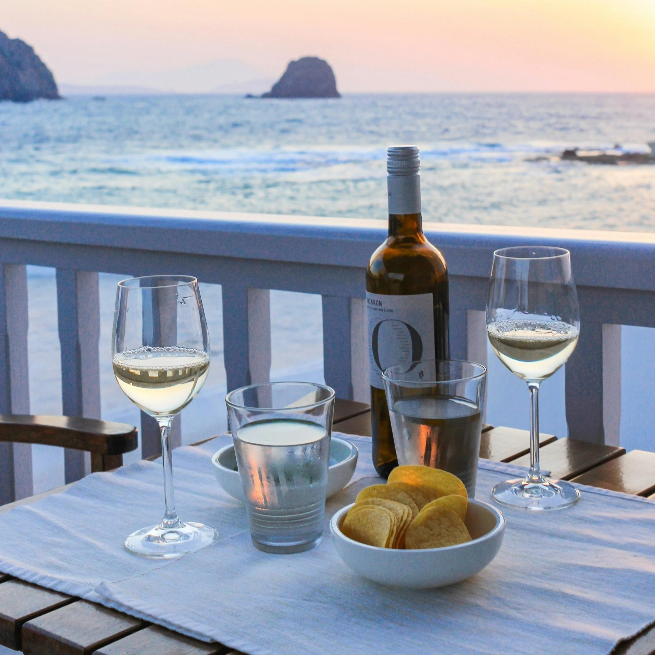 Mesa posta com duas taças de vinho branco, um copo de água, uma garrafa de vinho, uma tigela com salgadinhos e vista para o mar ao pôr do sol.