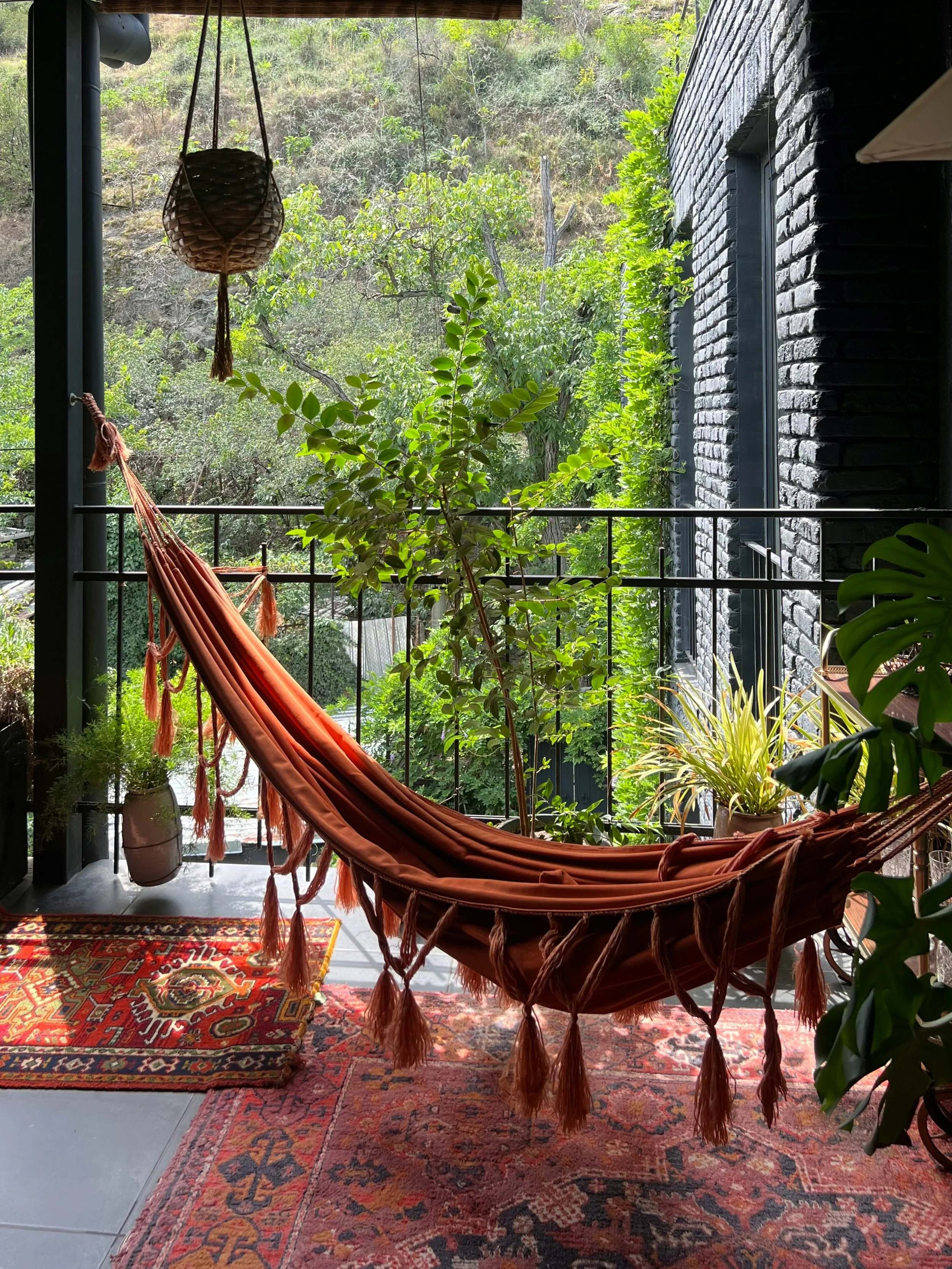 Varanda com rede de dormir cor de terracota, com tasséis, sobre tapetes coloridos, rodeada de diversas plantas verdes, com vista para uma área externa com árvores e vegetação.