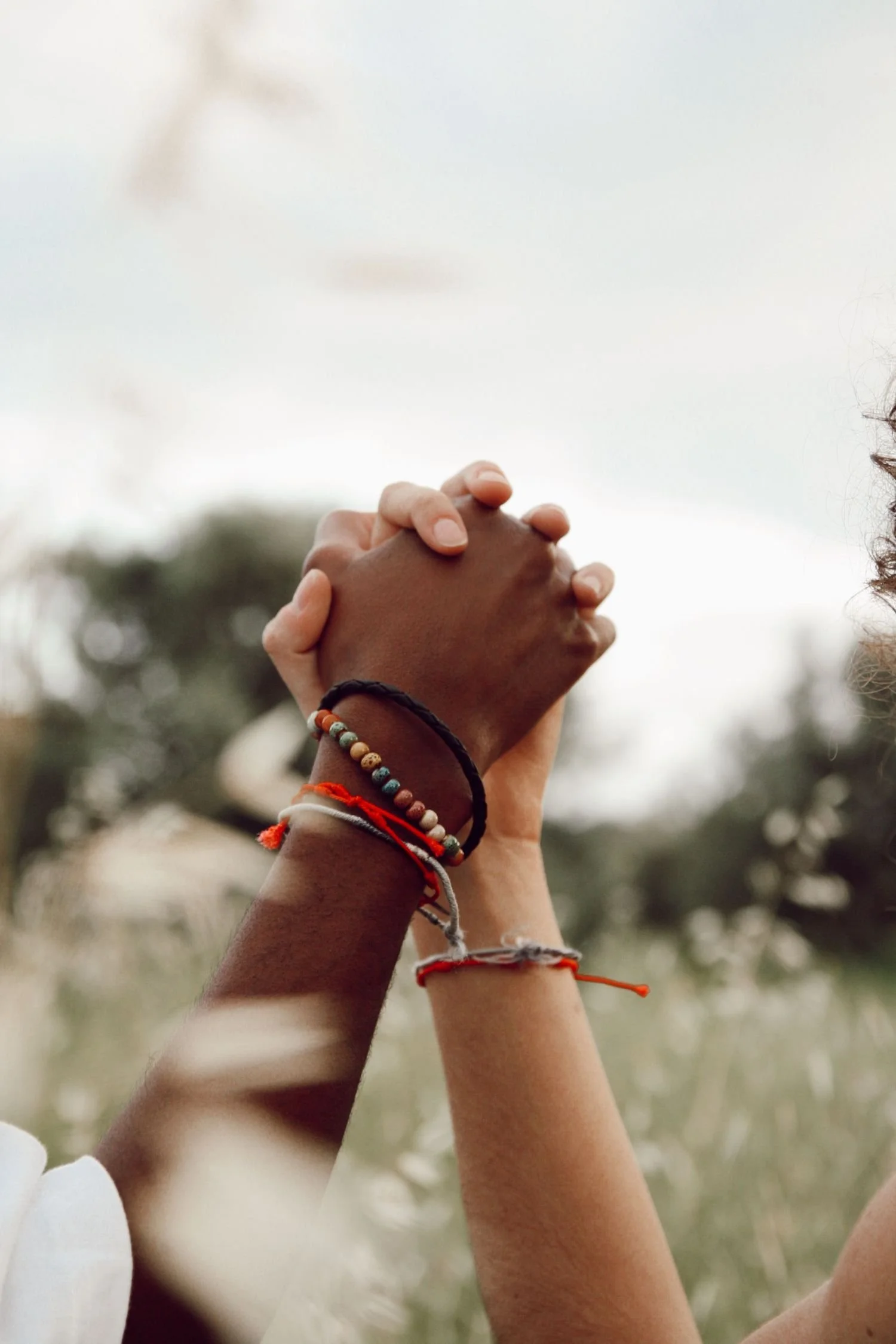 Two people holding hands, one with darker skin and the other with lighter skin, adorned with colorful bracelets, against a blurred outdoor background.