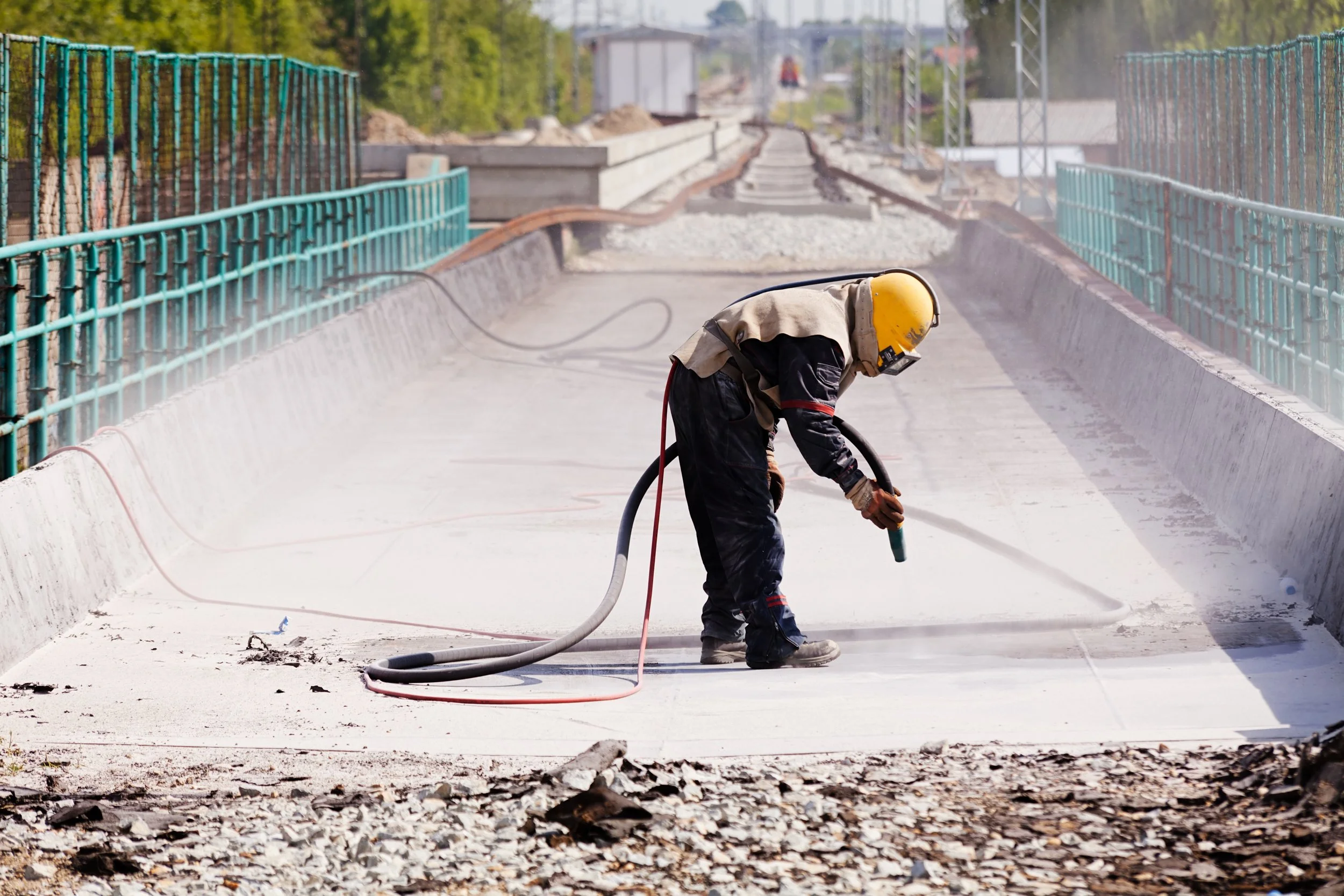 A construction worker wearing a yellow safety helmet and protective gear uses a power tool to grind a concrete surface on an outdoor railway path under construction, surrounded by safety fencing.