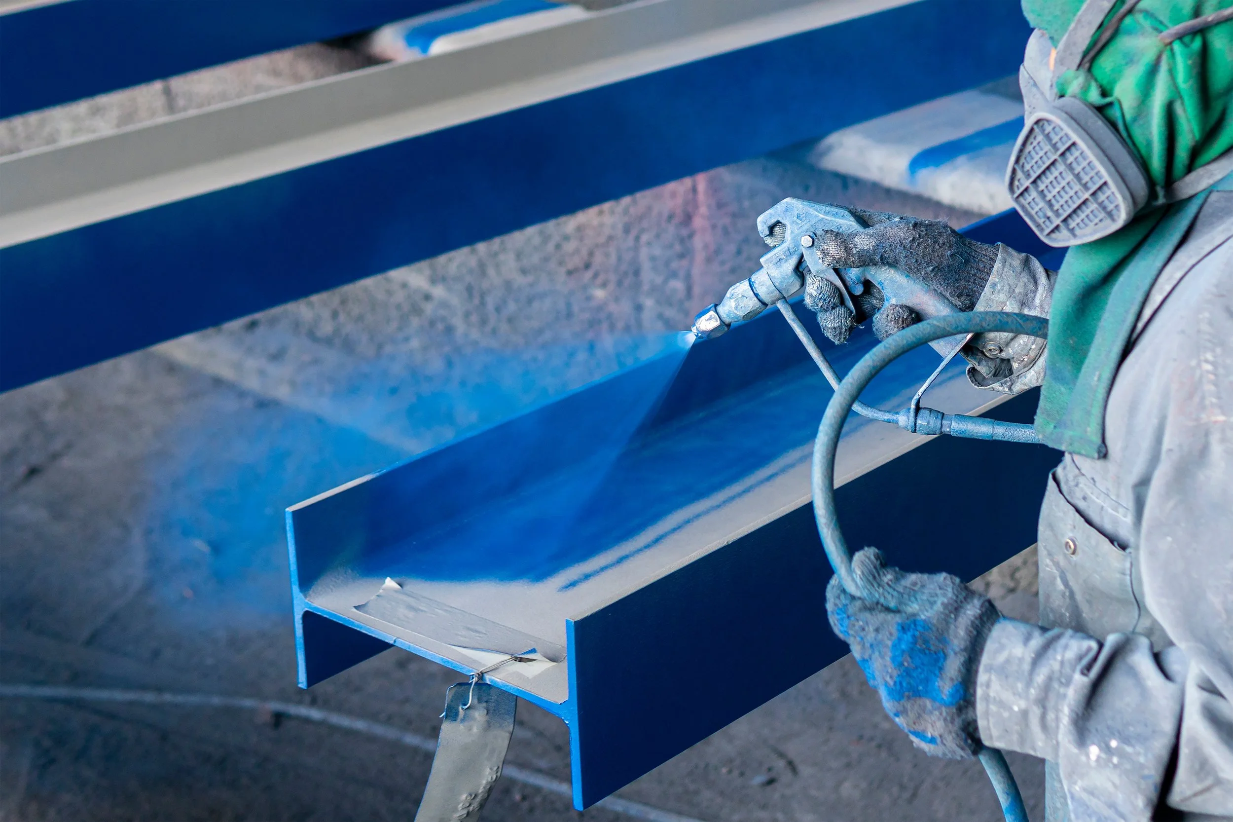 Worker wearing gloves and protective gear spray-painting a metal beam with blue paint in an industrial setting.