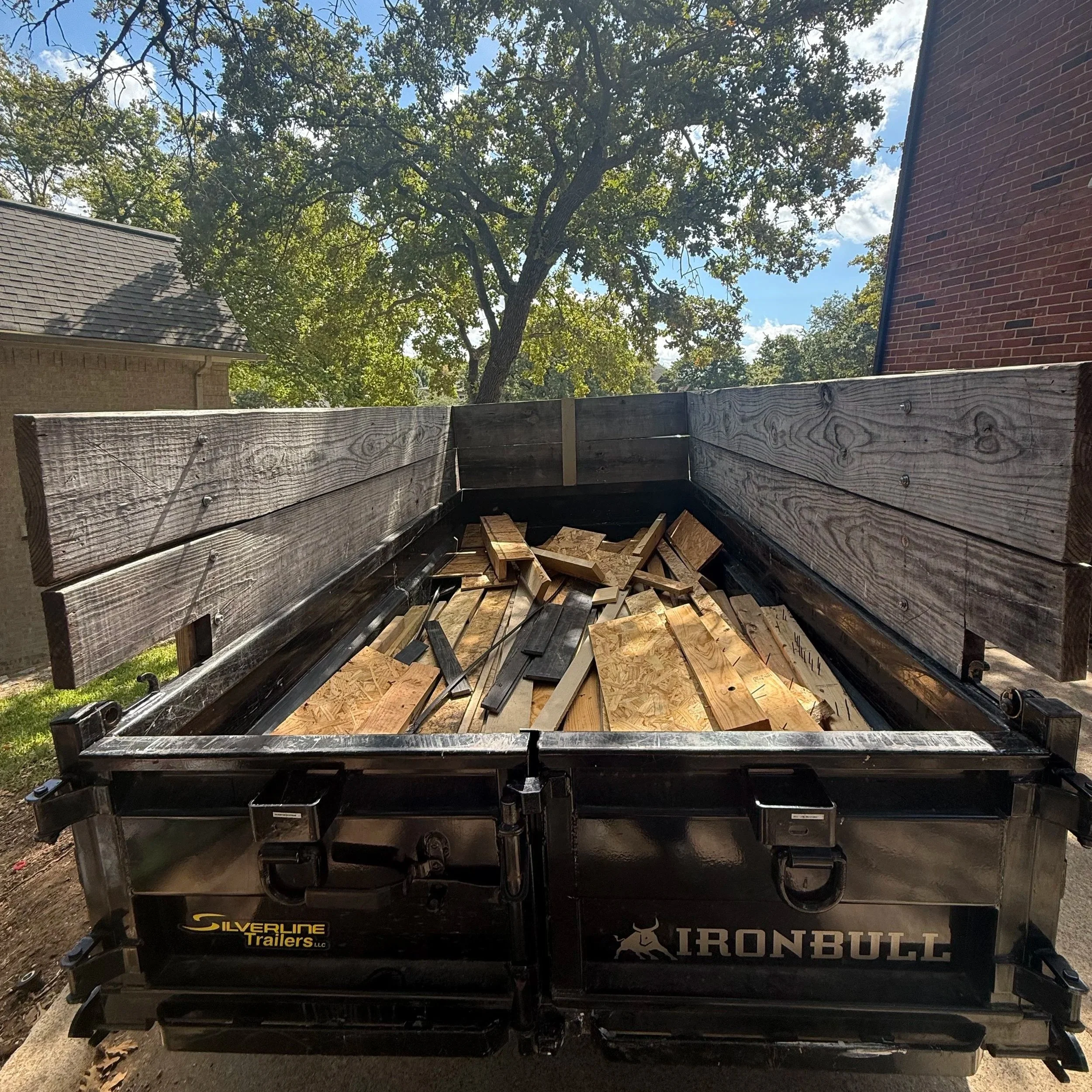 A large blue dumpster filled with construction or demolition debris, including wood, cardboard, fabric, and black plastic, in front of a gray multi-story house with siding and multiple windows.