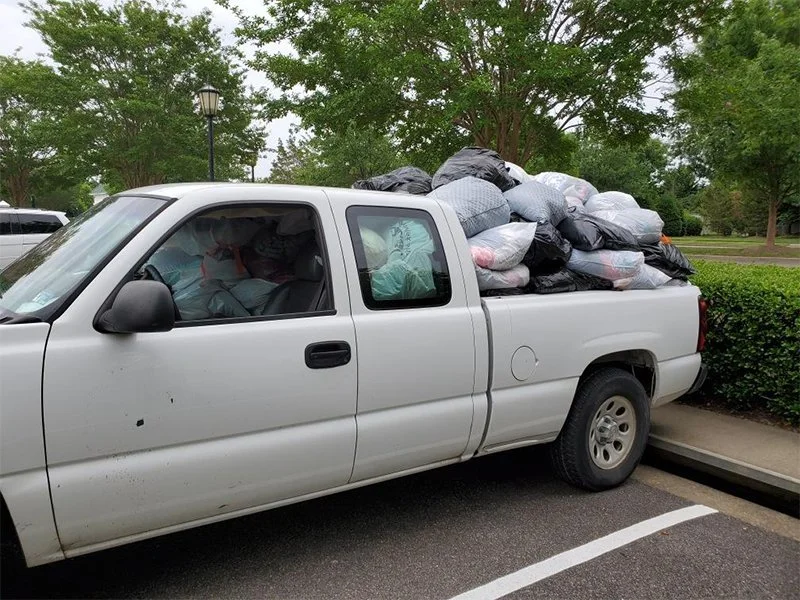 White pickup truck fully loaded with black and white trash bags in the bed, parked in a parking lot with trees and bushes in the background.
