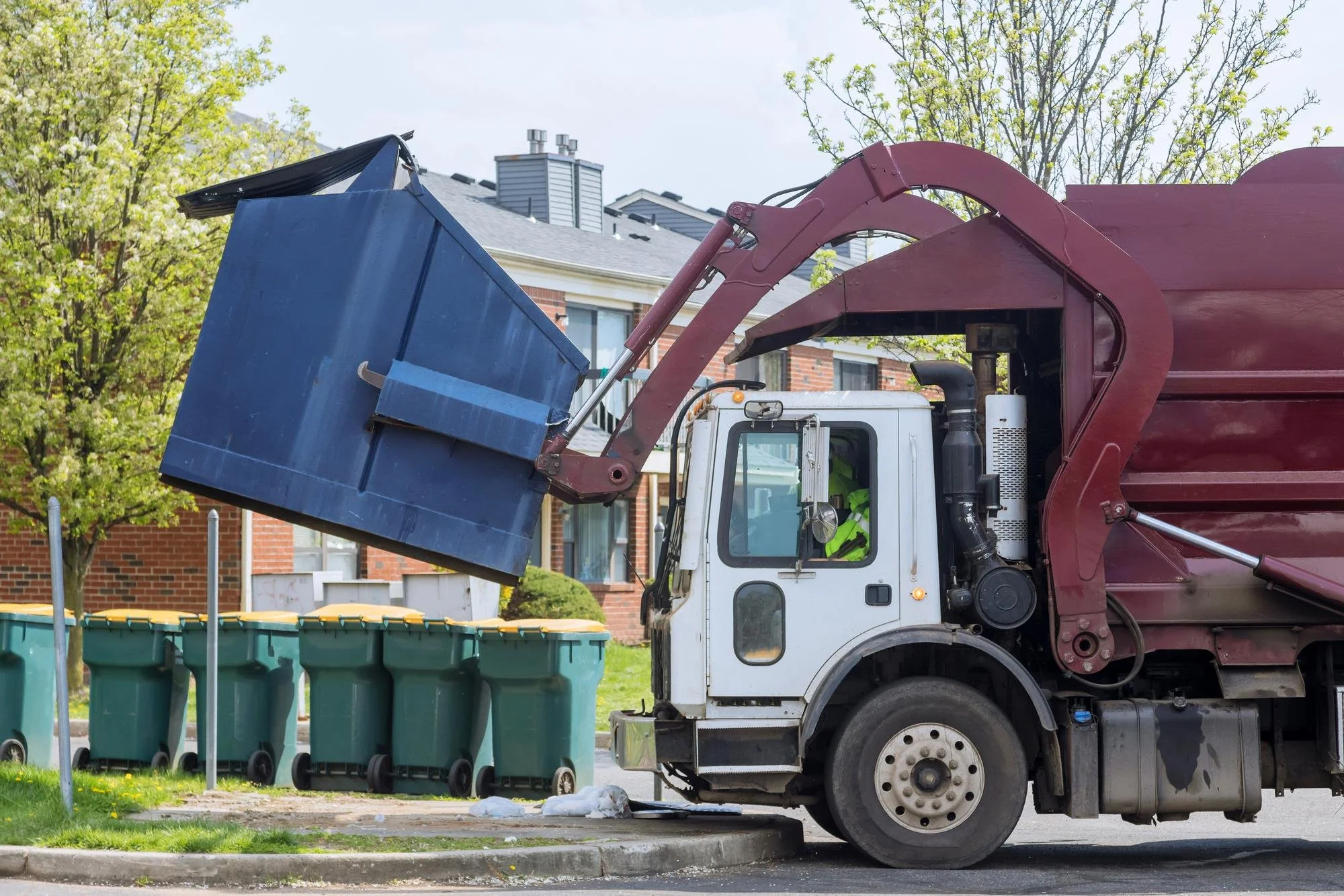 City street with garbage truck emptying trash bins, residential buildings, and trees in the background.