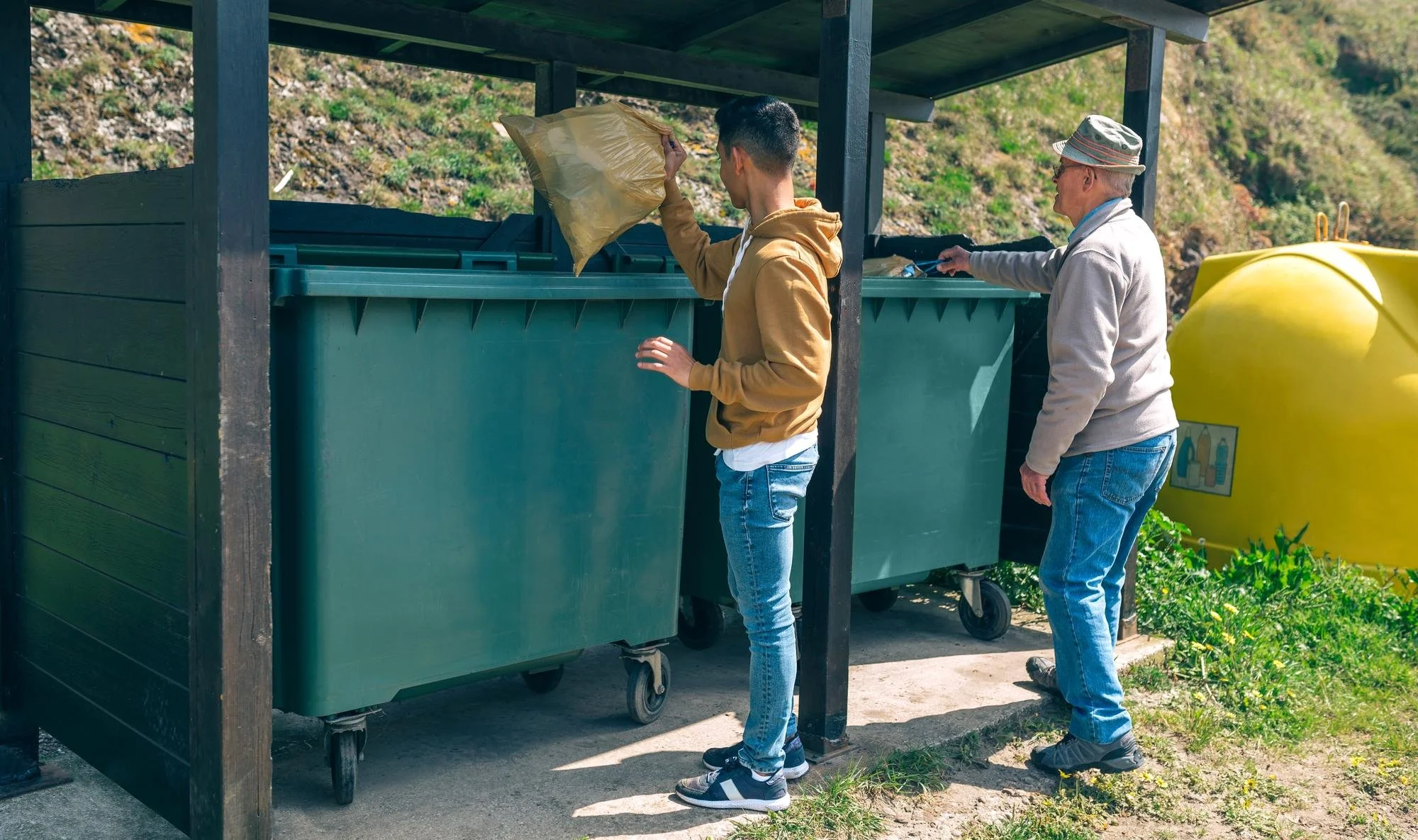 Two men recycling, one discarding a plastic bag into a green trash bin, and the other sorting items into another bin, outdoors near a yellow water tank and green plants.