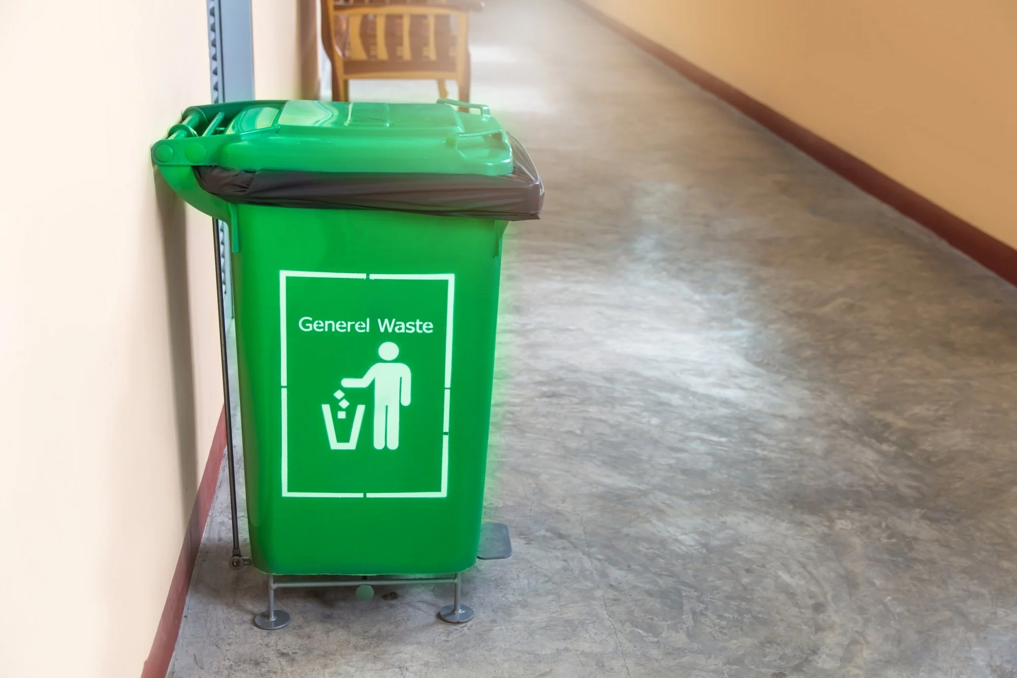 Green trash bin labeled 'General Waste' with a pictogram of a person disposing trash, placed against a beige wall in a corridor.
