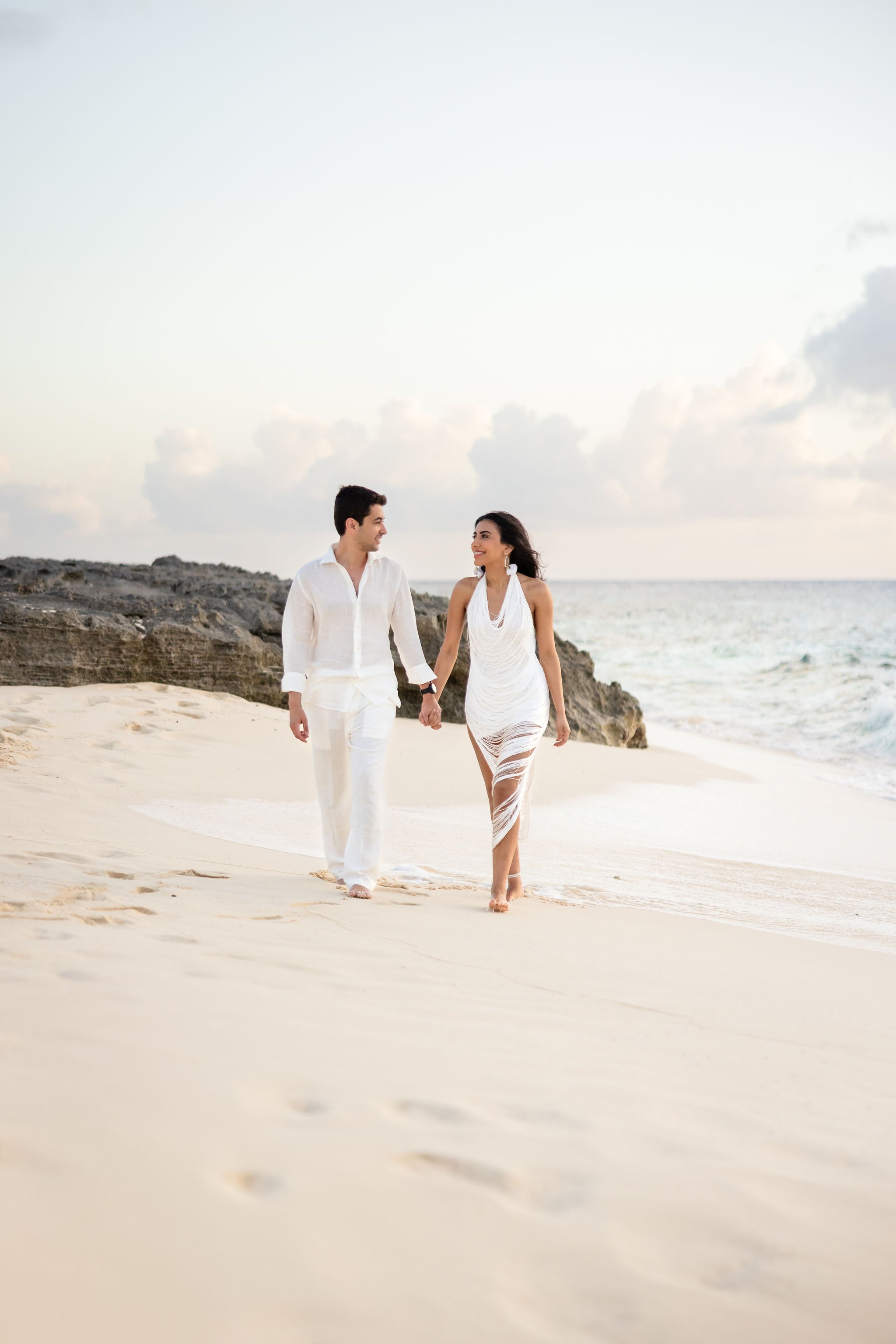 A couple dressed in white walking hand in hand along a sandy beach with rocks and ocean in the background at sunset.