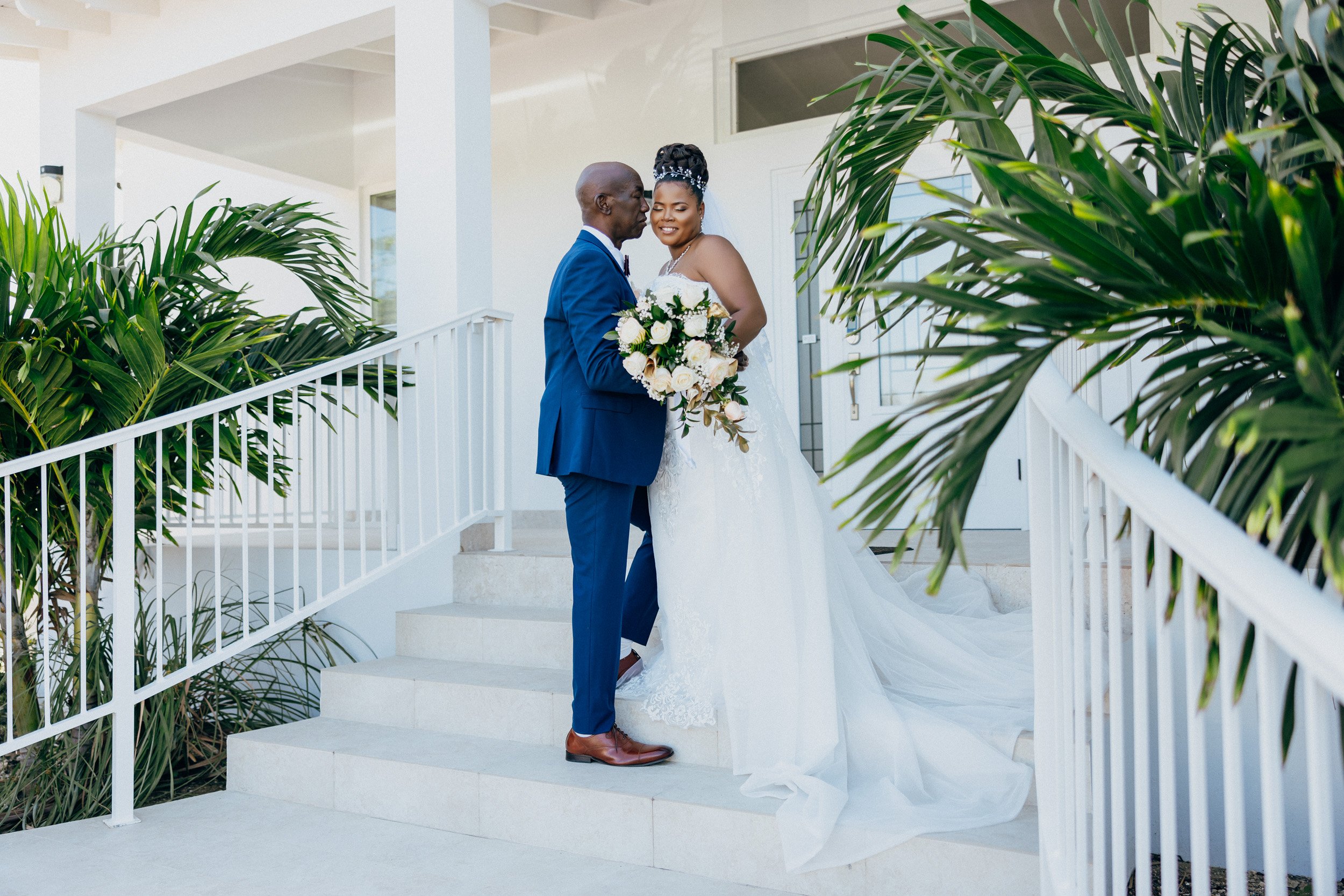 A bride and groom standing on white stairs outside a white building, sharing a tender moment with the groom gently kissing the bride's forehead. The bride is holding a bouquet of white and green flowers, wearing a white wedding gown, and has a bejewe
