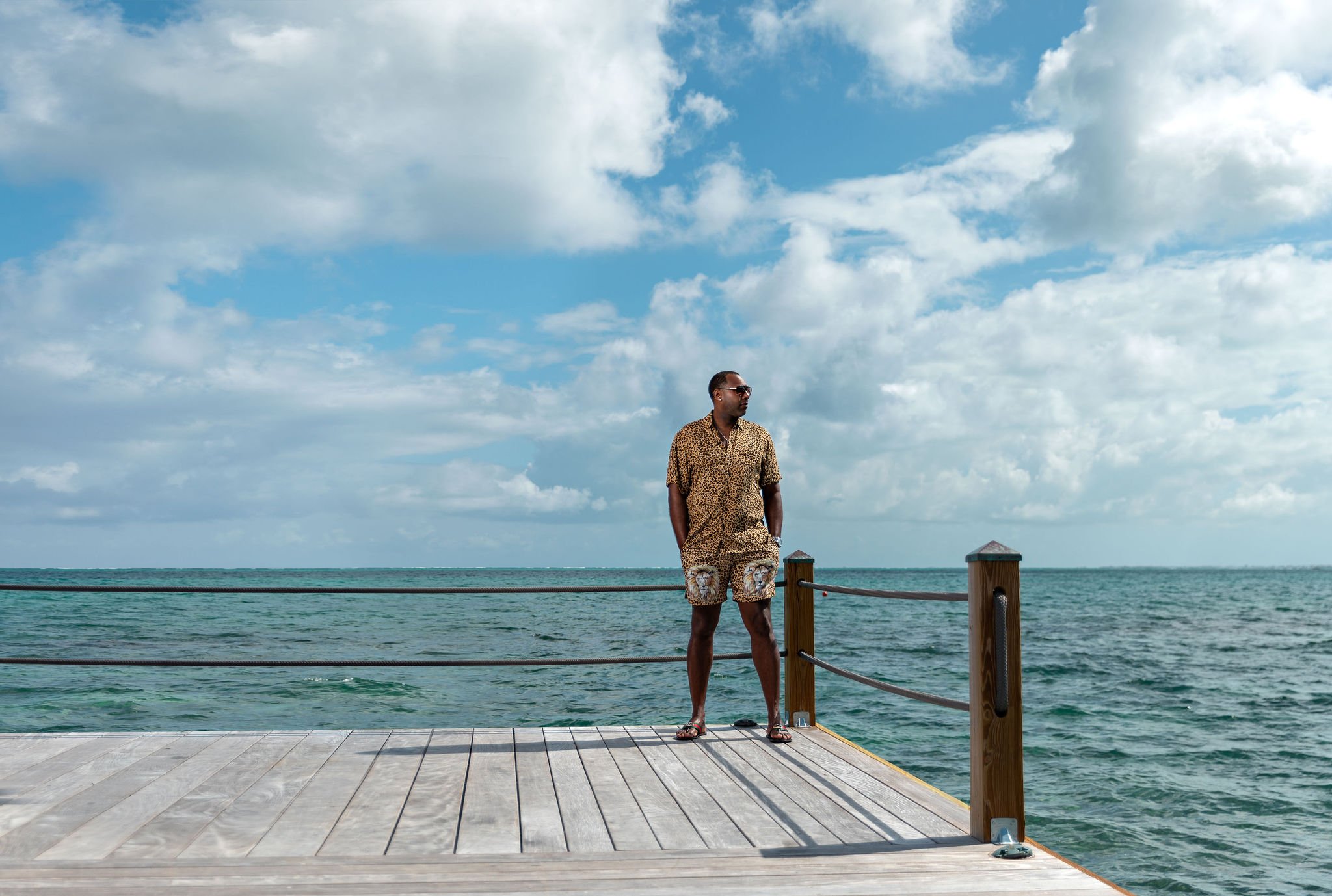 A man in leopard print clothing standing on a wooden dock over the ocean with a partly cloudy sky in the background.