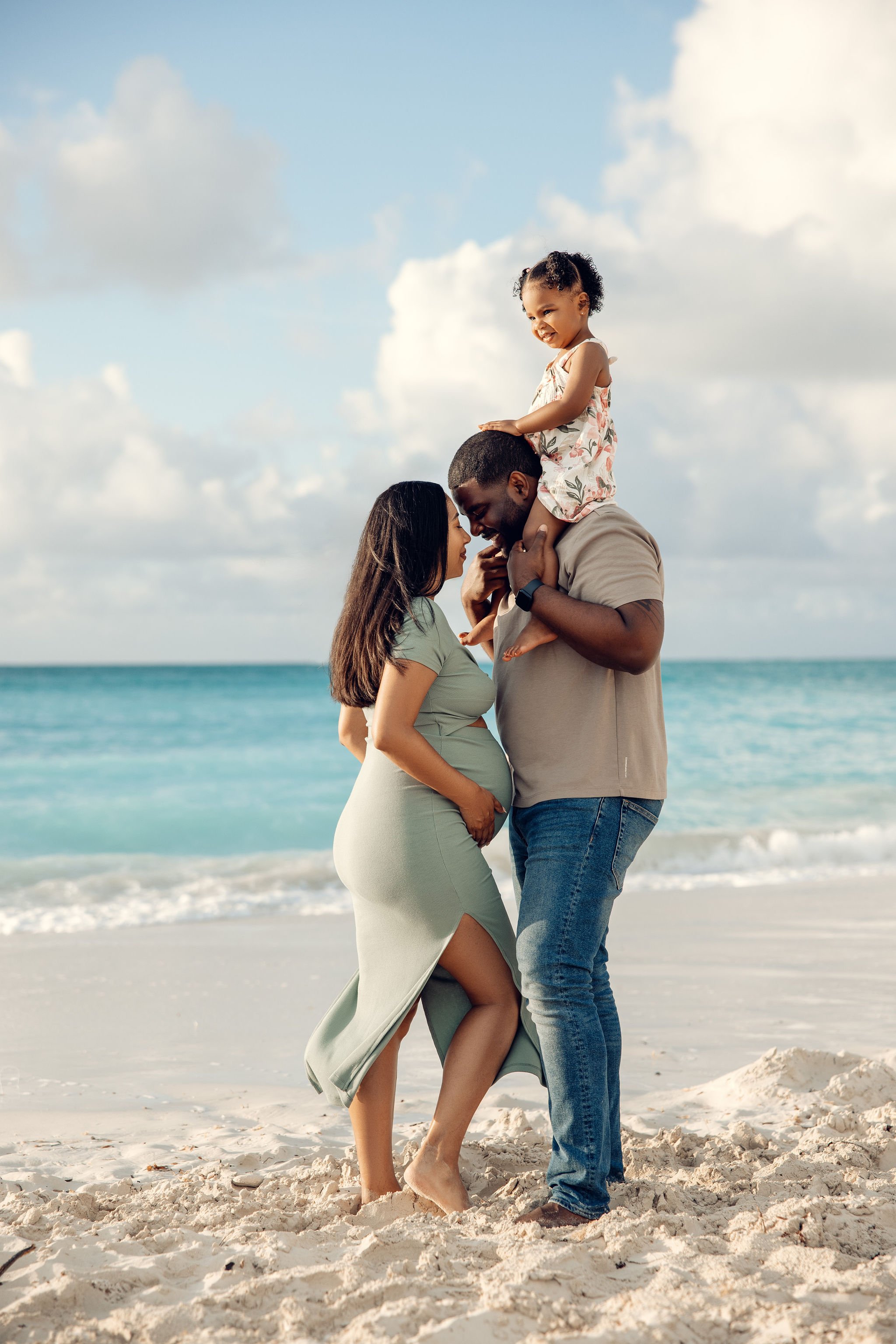 A family of three, including a pregnant woman, a man, and a young girl, enjoying a moment together on a sandy beach with ocean and sky in the background.