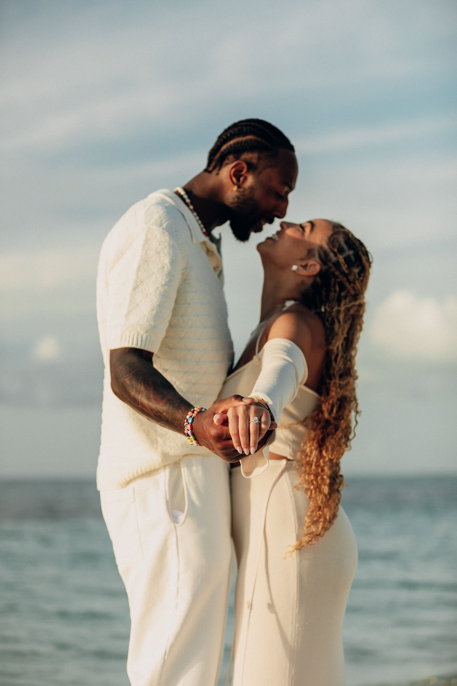A couple holding hands and smiling at each other on the beach during sunset.
