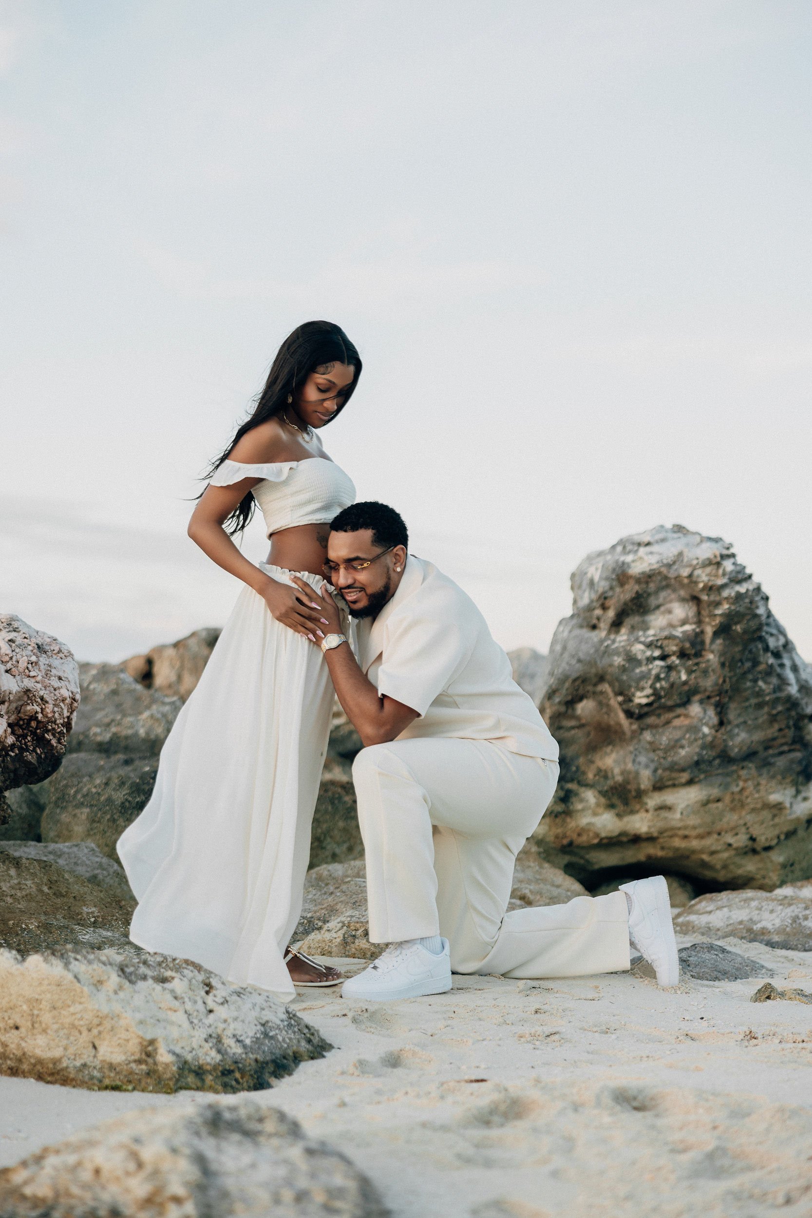 A pregnant woman wearing a white crop top and skirt standing on rocks at the beach, with a man kneeling in front of her holding her belly and smiling.