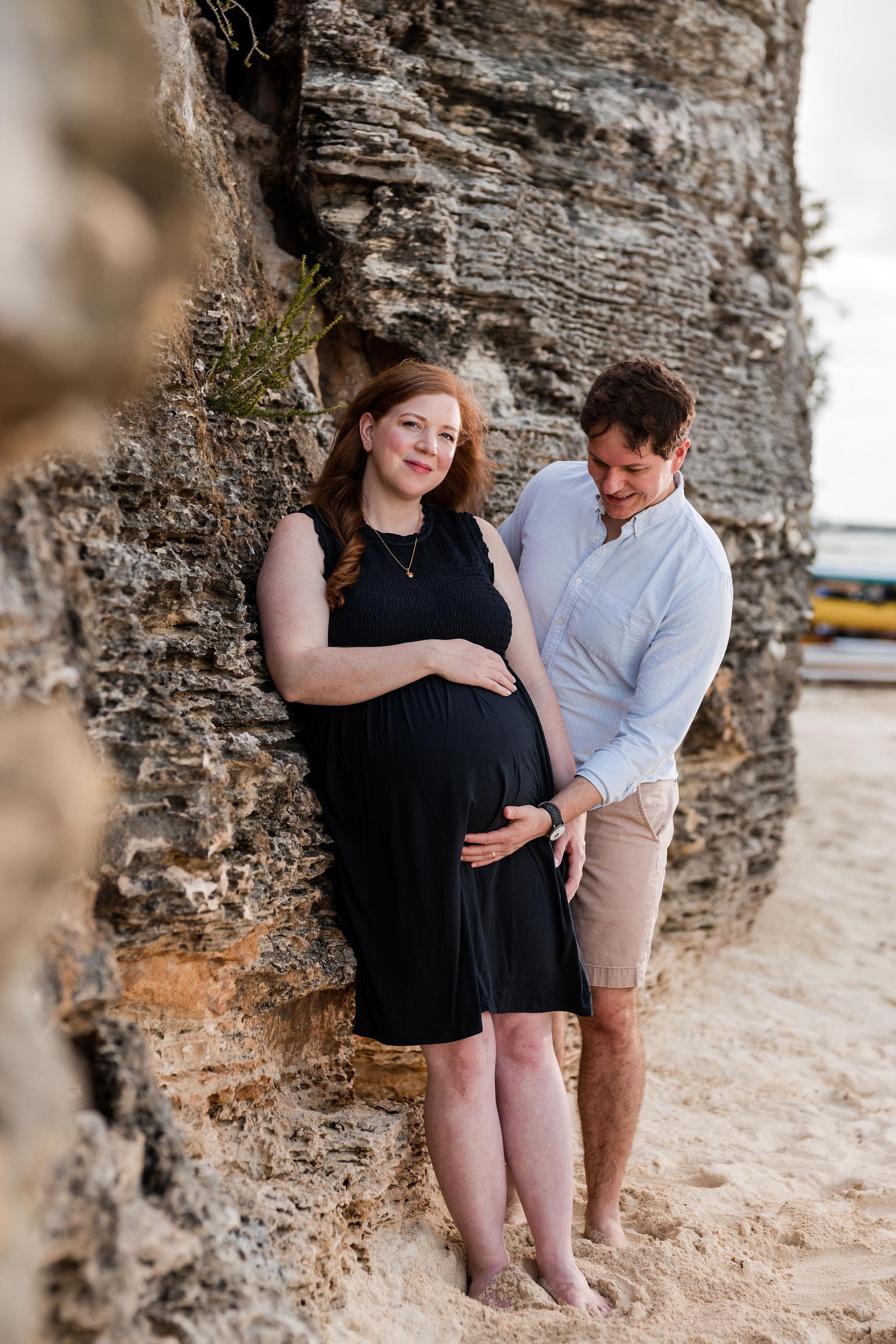 Pregnant woman in a black dress leaning against a rocky wall on the beach, with a man standing behind her holding her belly and smiling.
