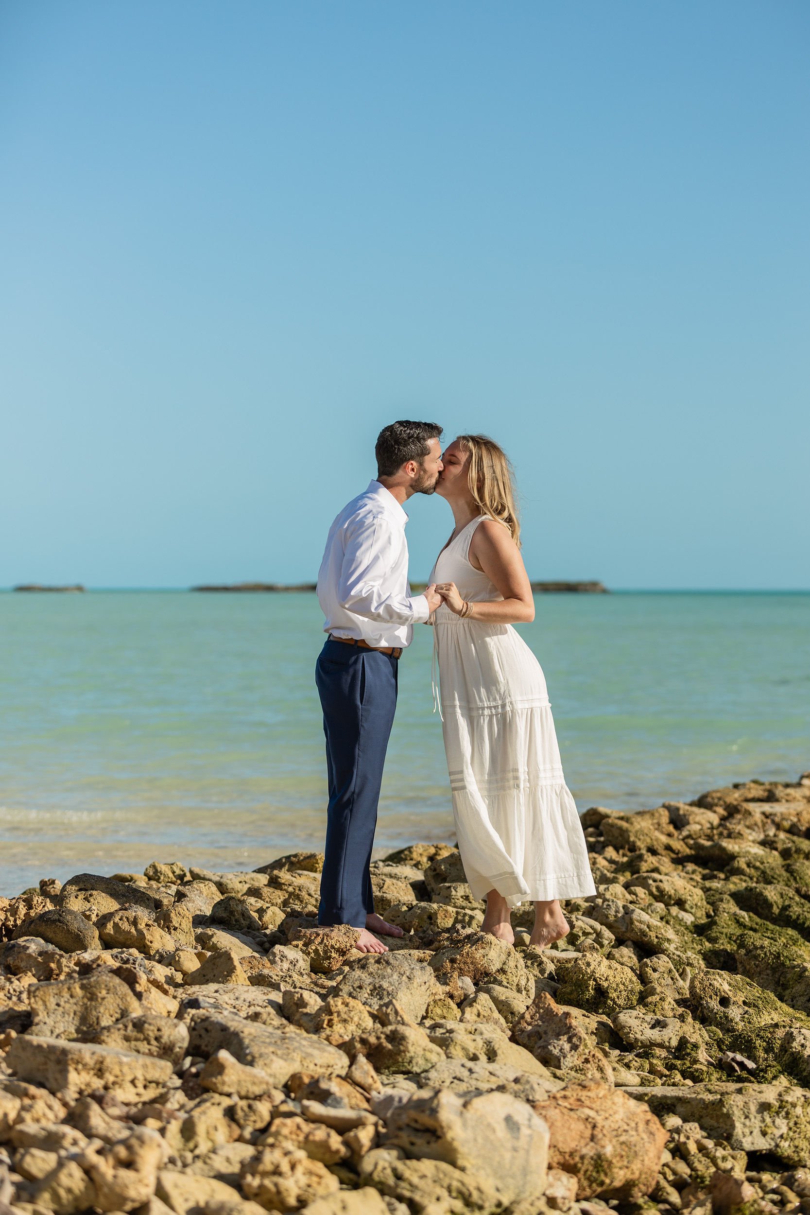 A couple is standing on a rocky shoreline, kissing, with ocean water and a clear blue sky in the background.
