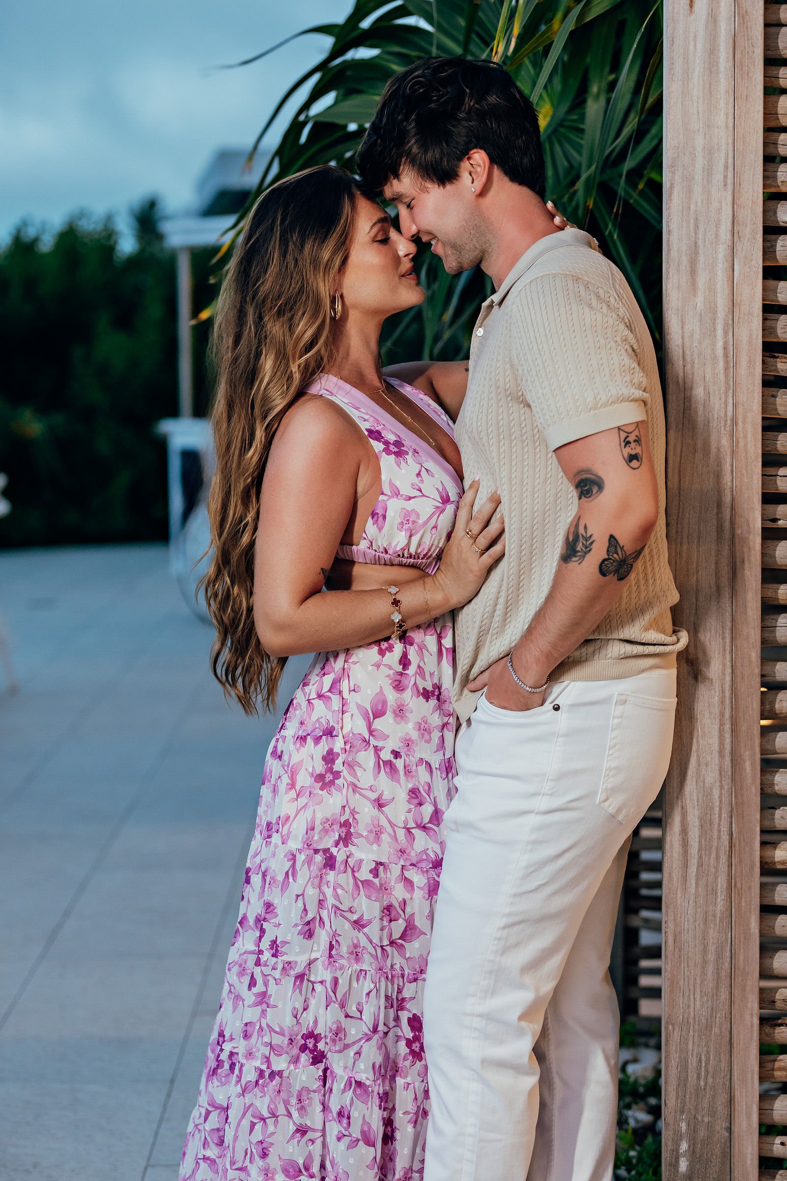 A man and woman are standing close with their foreheads touching, embracing each other. The woman has long wavy hair and wears a pink floral dress. The man has short dark hair, tattoos on his arm, and is dressed in a beige polo shirt and white pants,