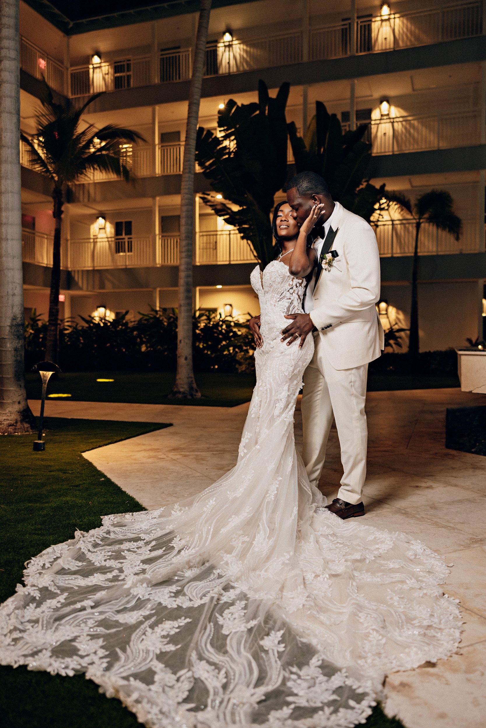 A bride and groom dancing outdoors at night, with palm trees and a multi-story building in the background.