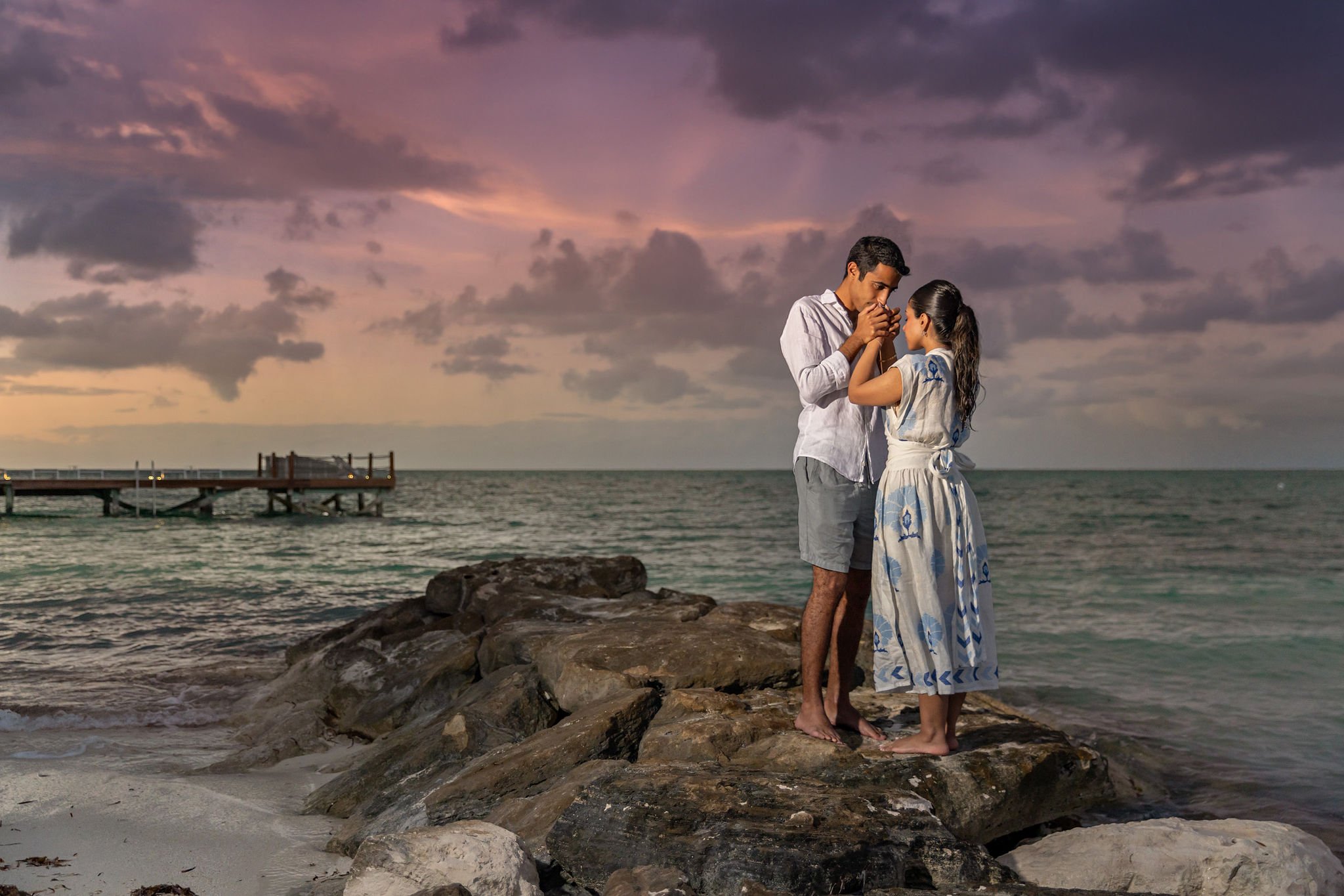 A couple stands on a rock by the beach during a sunset, holding hands and gazing at each other, with a pier in the background and a cloudy sky in hues of pink and purple.