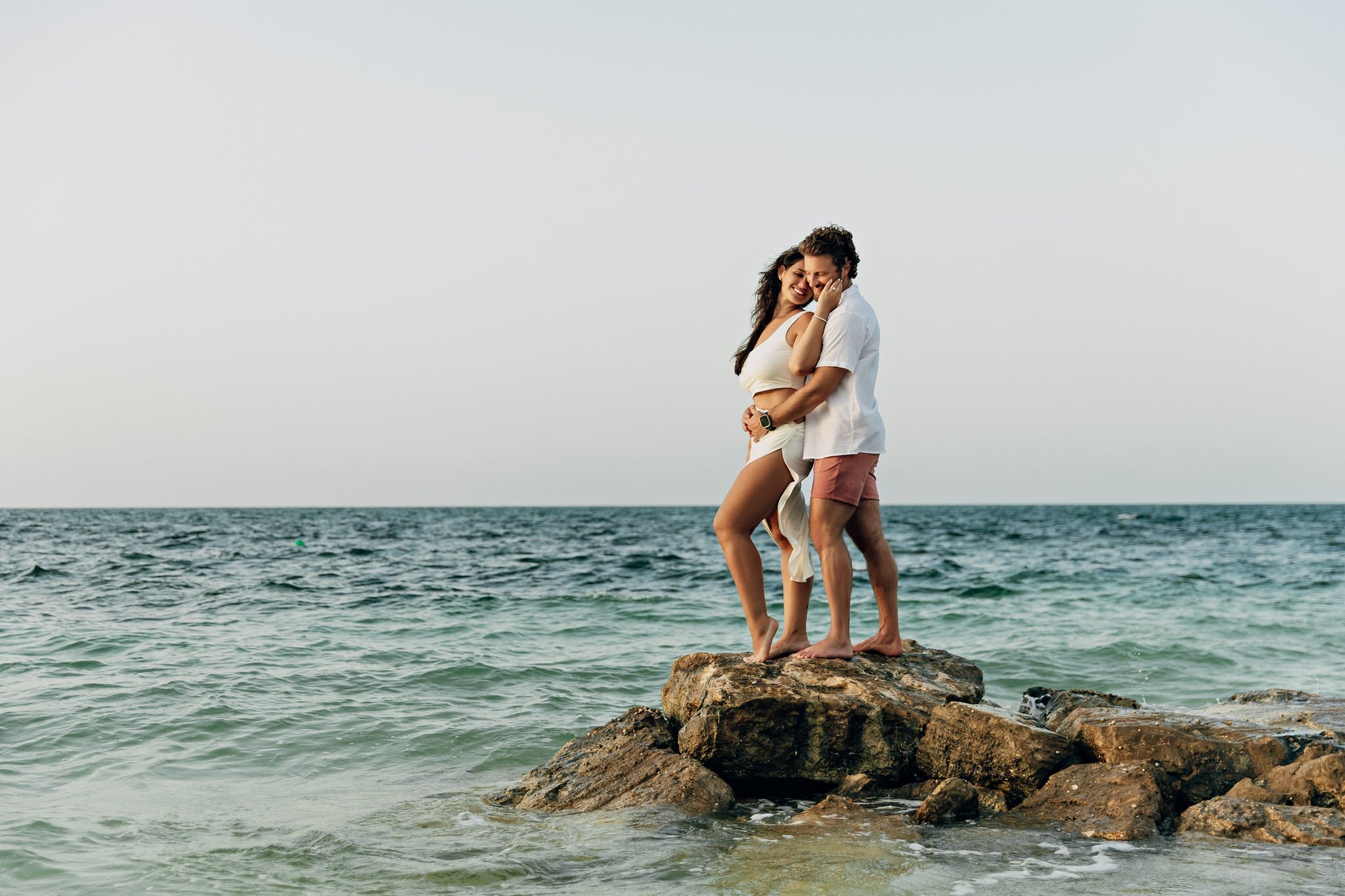 A couple standing on rocks at the beach, smiling and embracing, with ocean waves in the background under a cloudy sky.