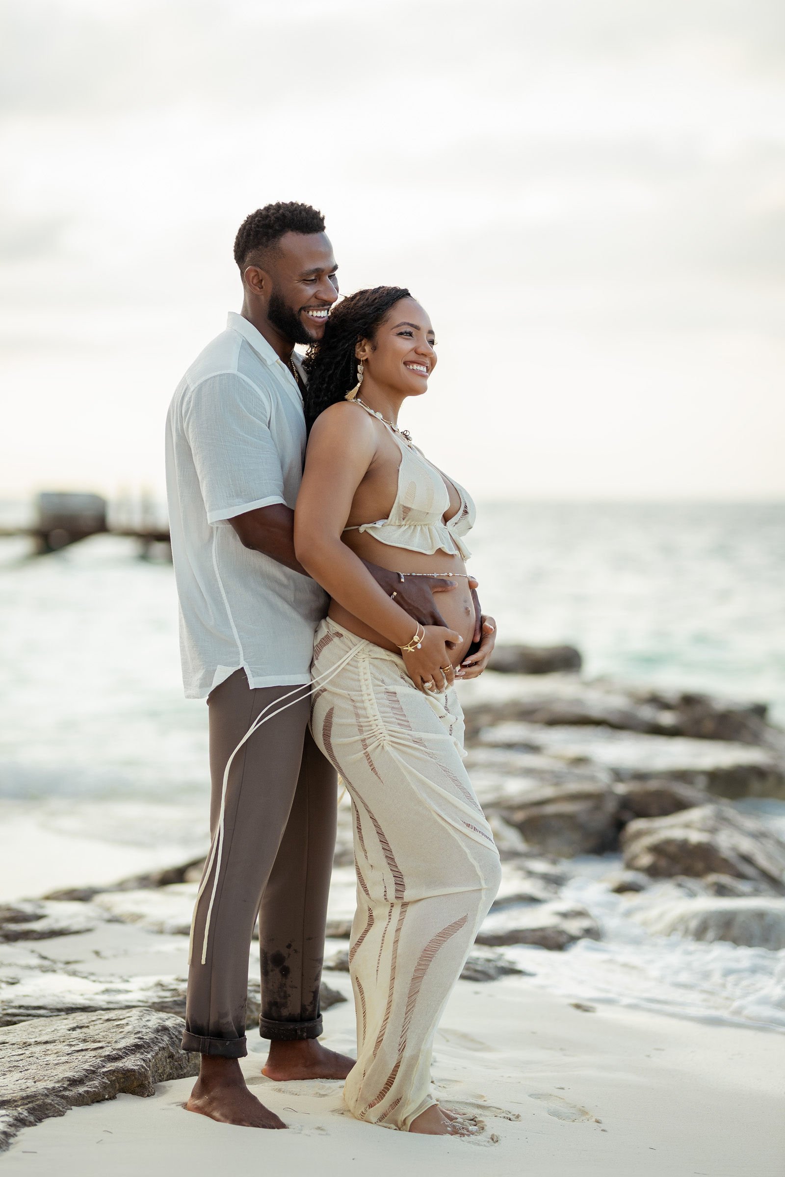 A happy couple standing on a beach, with the man gently holding the pregnant woman's belly. They are smiling and looking joyful, with the ocean and rocks in the background.