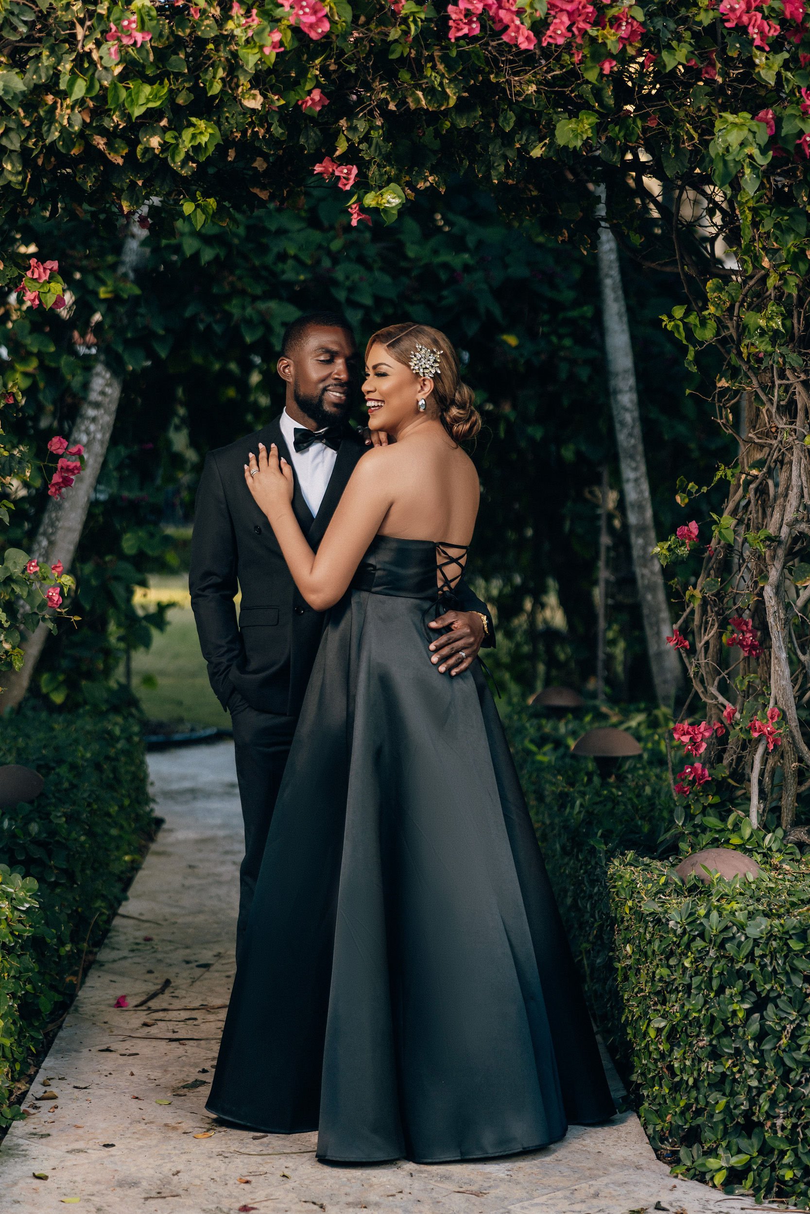 A couple in formal attire, a man in a tuxedo and a woman in a black strapless gown, are embracing outdoors under a floral archway with pink flowers and green foliage.